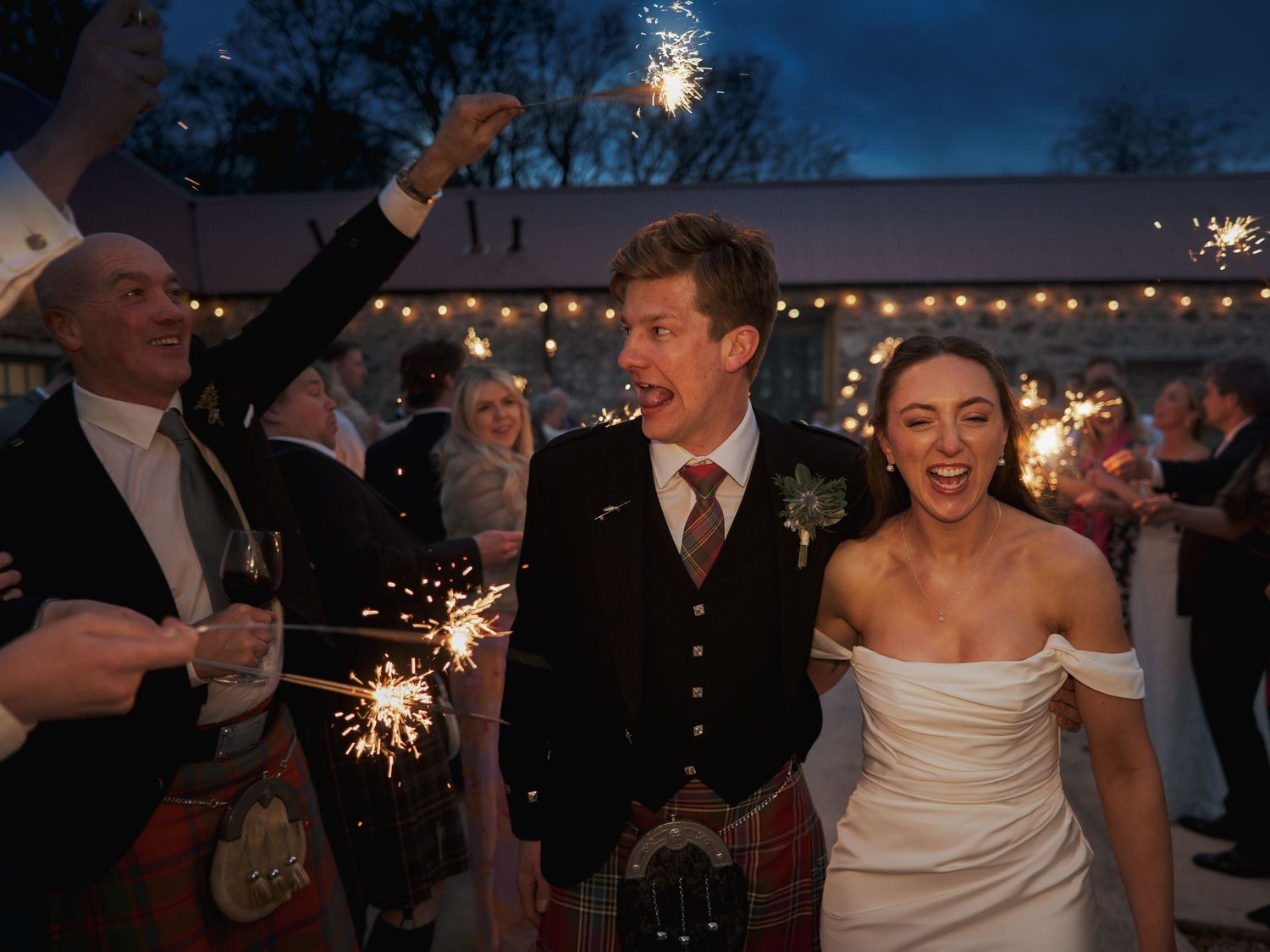 Bride and groom walk through sparkler send-off at Scottish wedding, groom wearing tartan kilt.