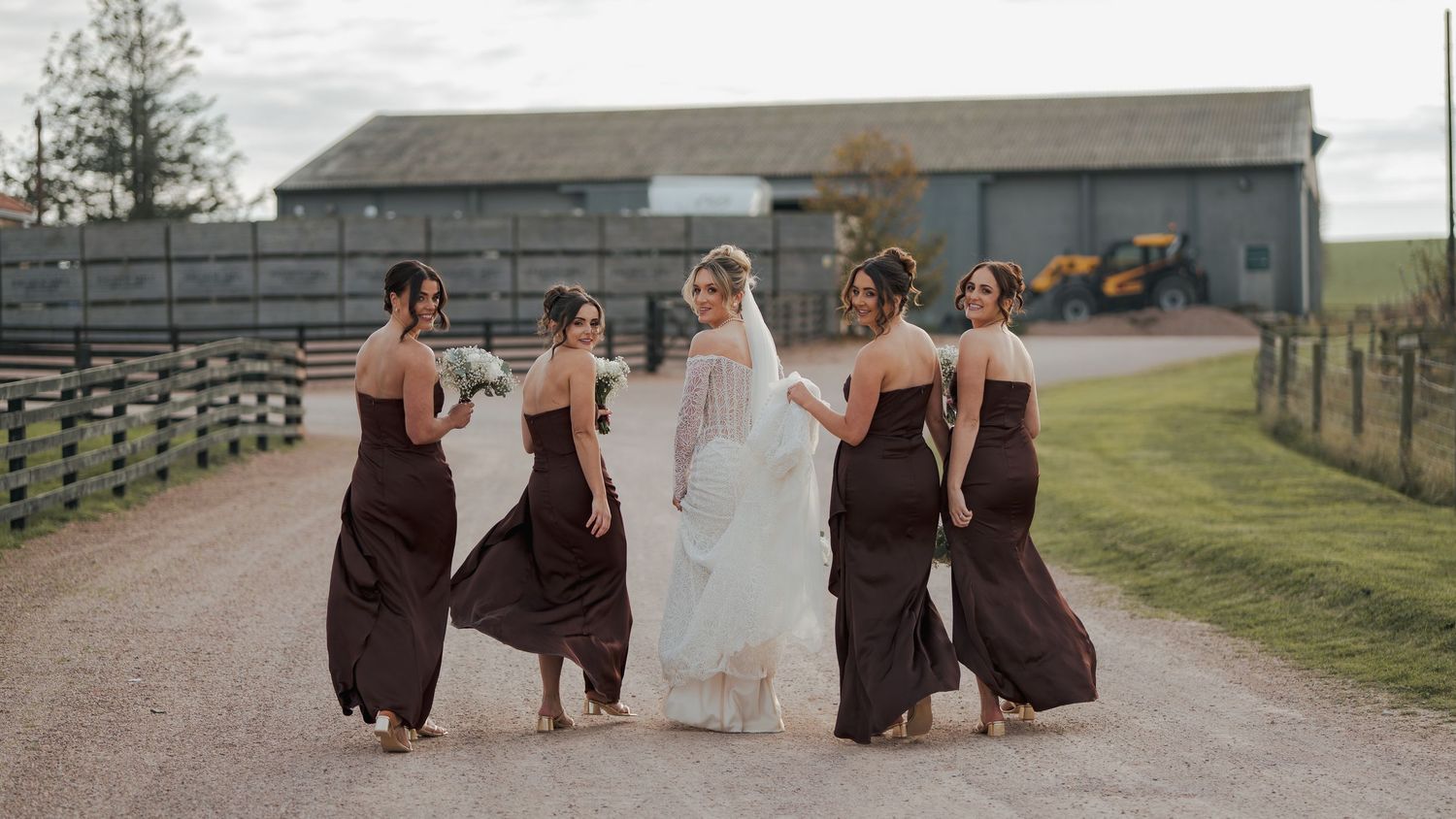 A group of bridesmaids in brown dresses walk down a gravel path with the bride at Falside Mill.