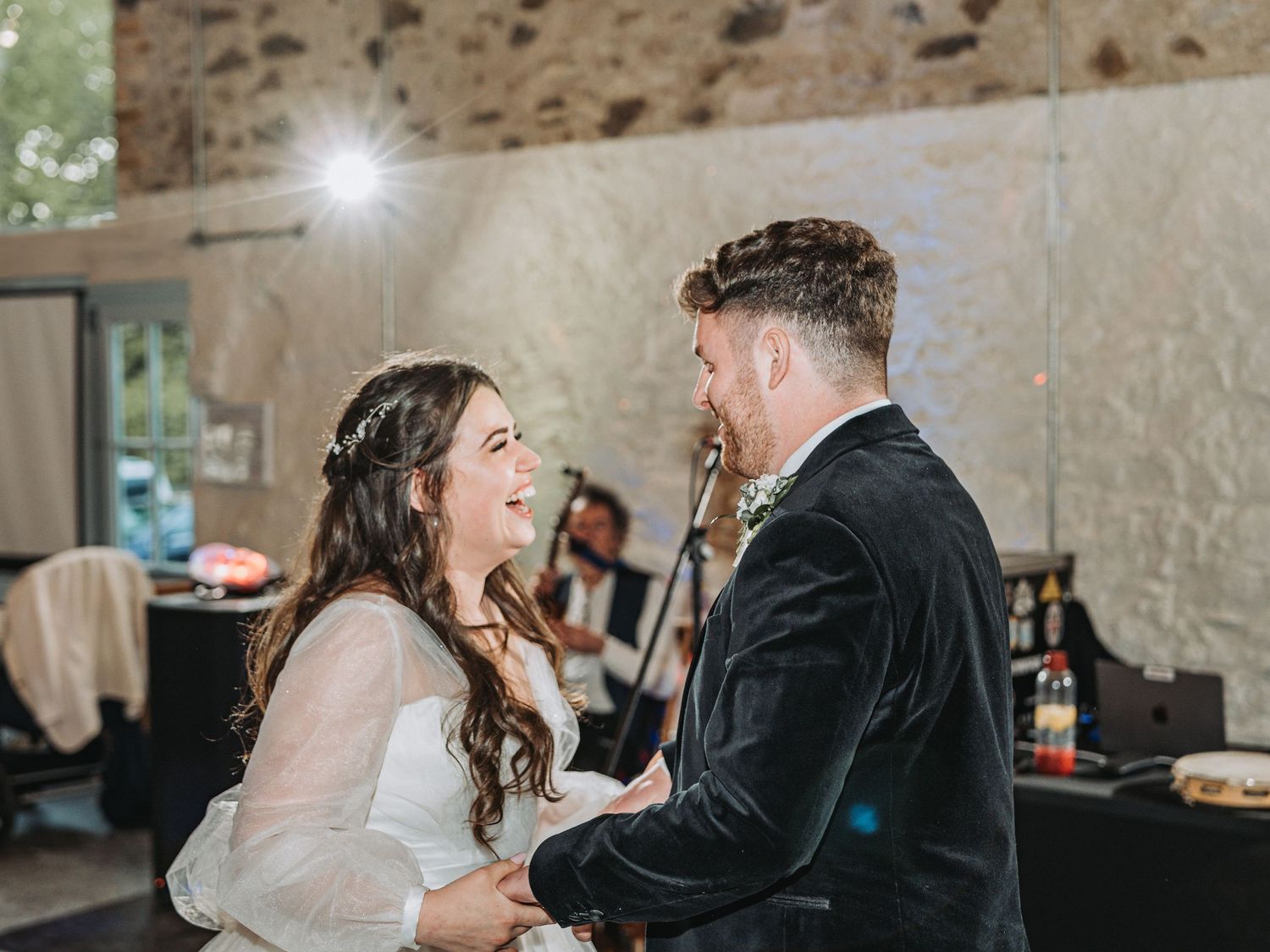 Bride and groom share a joyful candid moment dancing together at their wedding reception in a rustic stone venue.