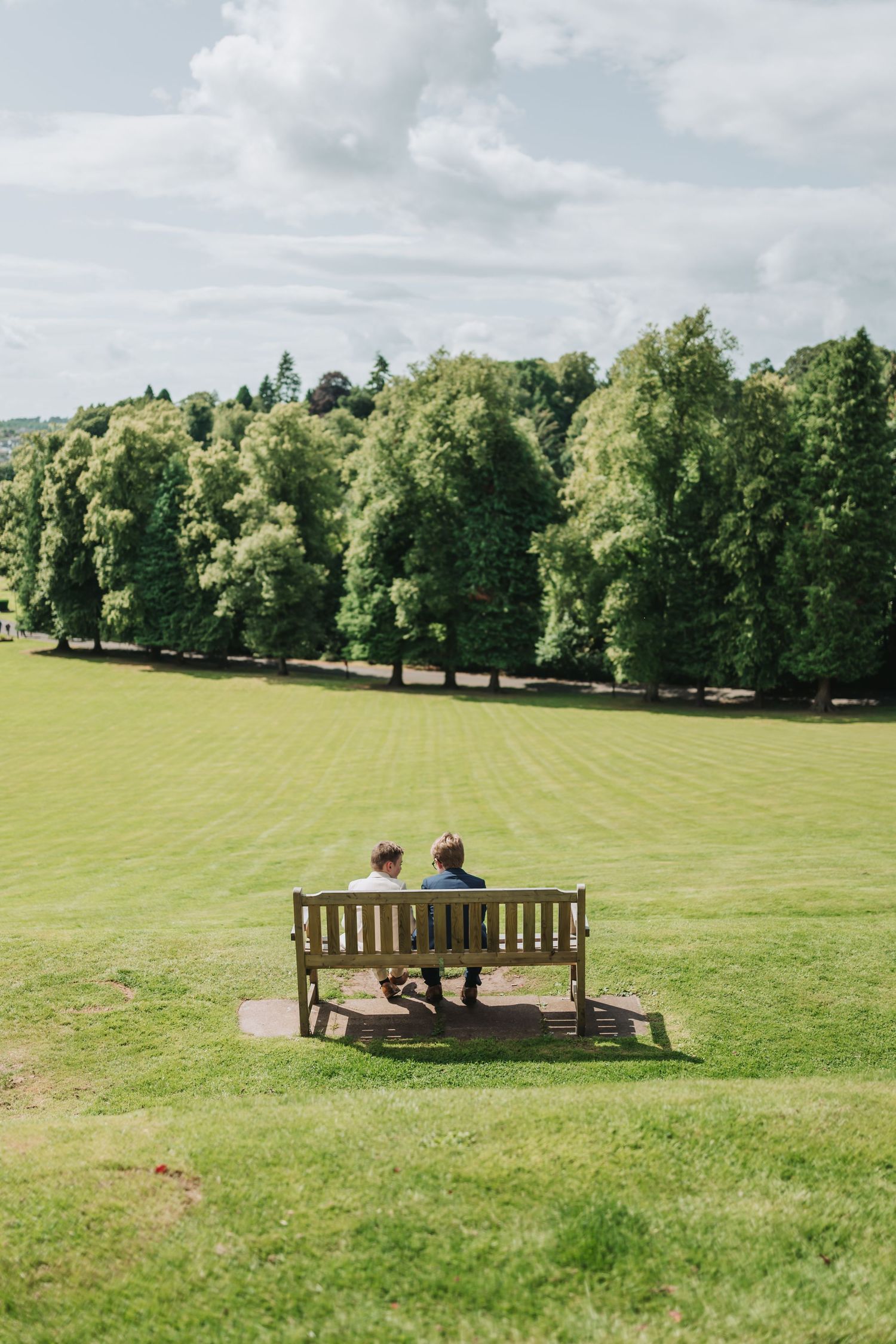 Two people sitting on a wooden bench at Dunblane Hydro overlooking a serene green lawn bordered by tall trees on a cloudy summer day.