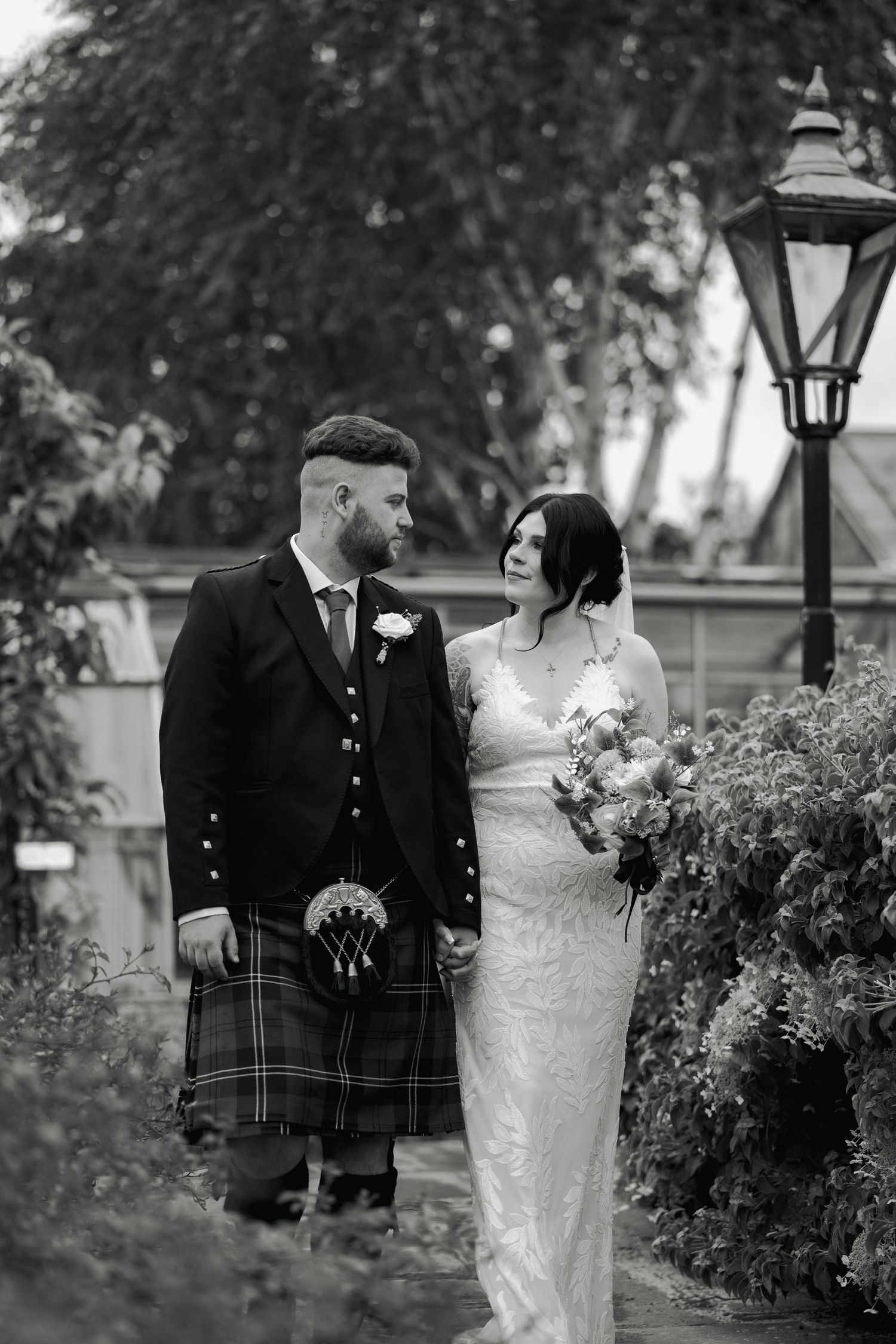 Black and white photo of a bride in lace gown and groom in Scottish kilt holding hands in a garden setting.