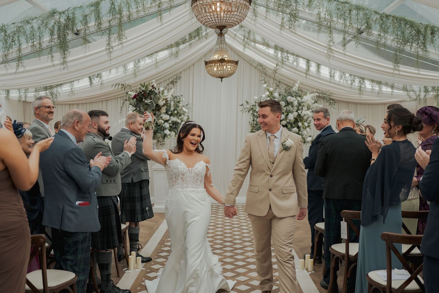 Bride and groom walk down the aisle holding hands in an elegant marquee wedding venue with floral decor.