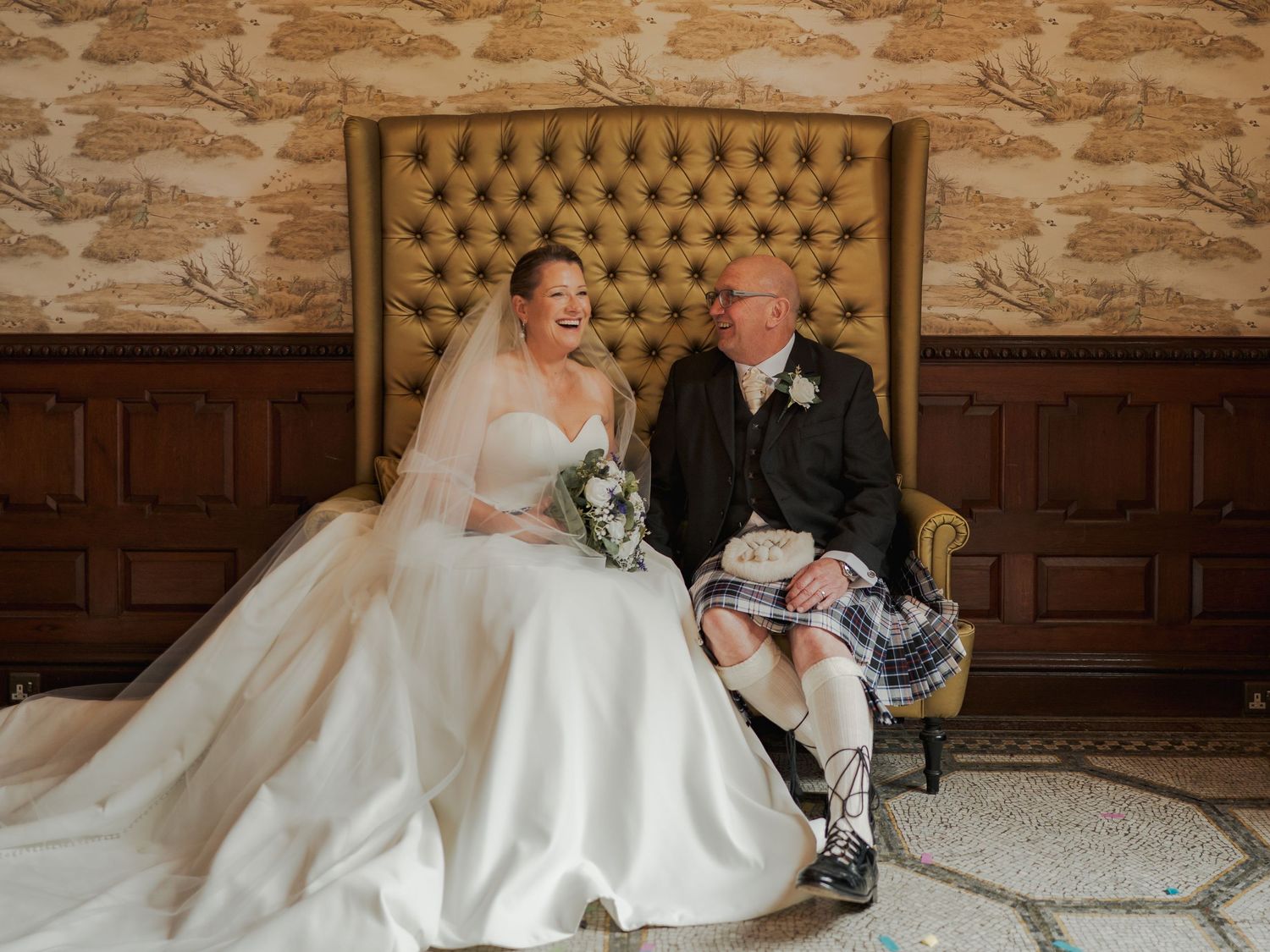 Bride and groom laughing together on a tufted gold sofa, wearing wedding dress and Scottish kilt.