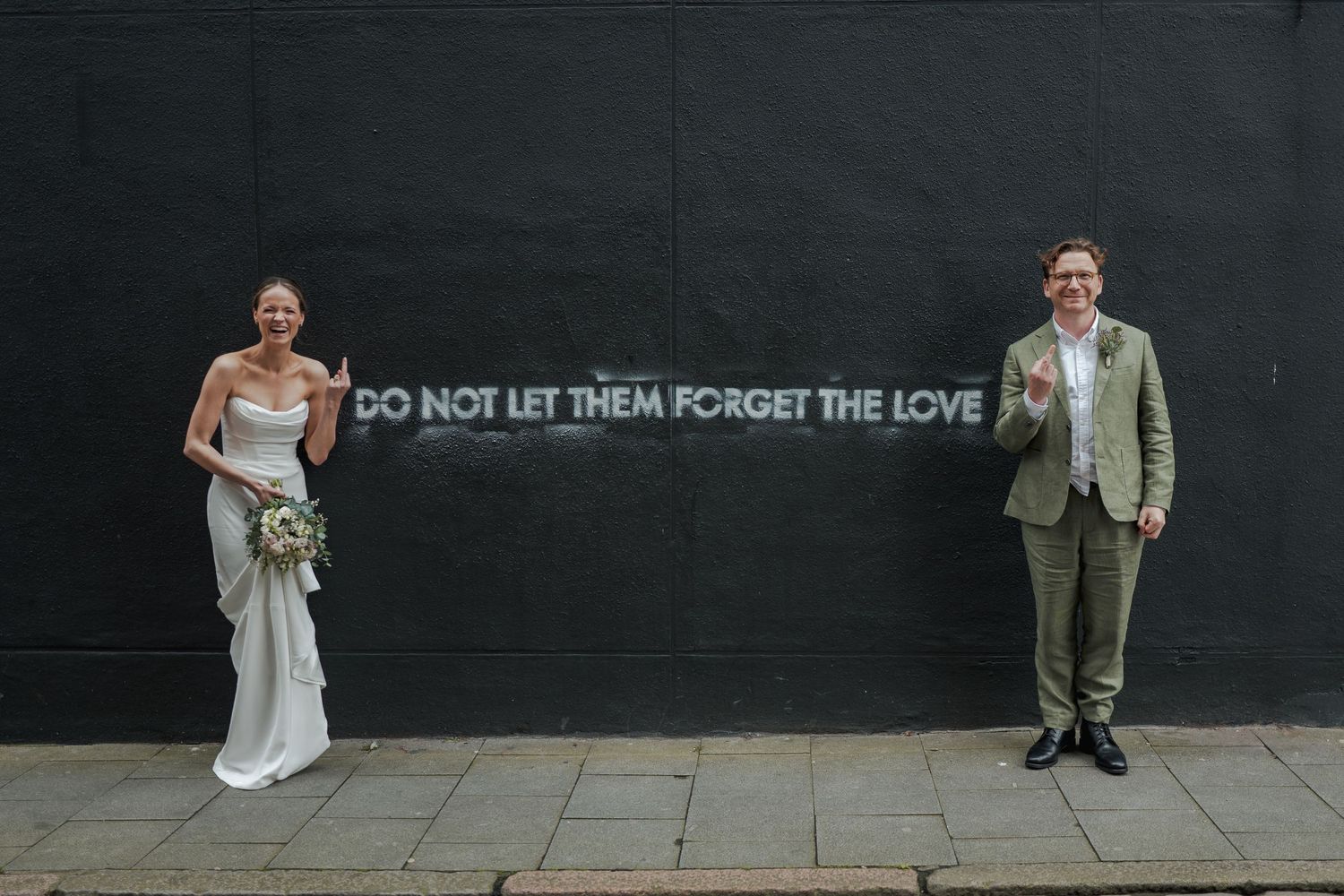 Bride and groom posing apart beside a black wall with neon text reading Do Not Let Them Forget The Love.