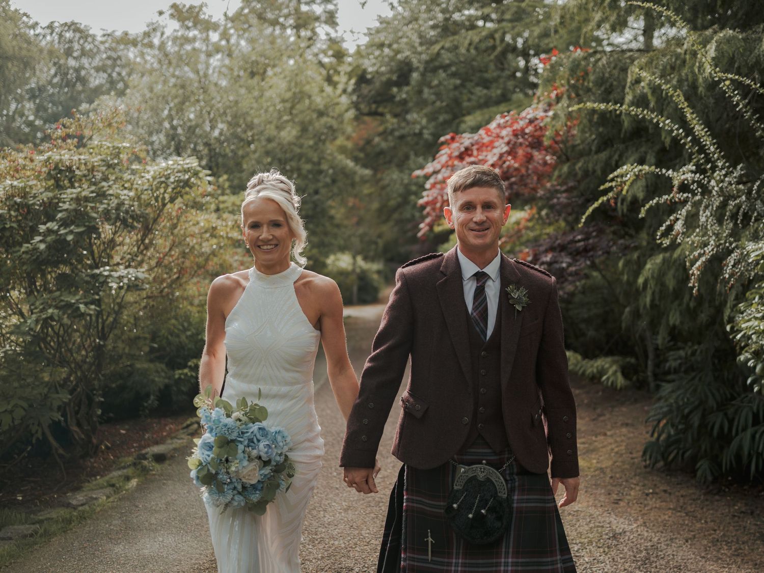 Bride in white halter gown and groom in traditional Scottish tartan kilt walk hand in hand through lush garden path.