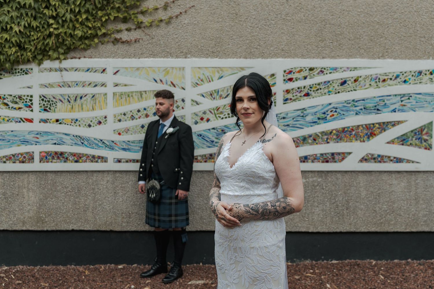 Bride in lace wedding dress holding clutch stands before mosaic wall as groom in Scottish kilt stands behind her.