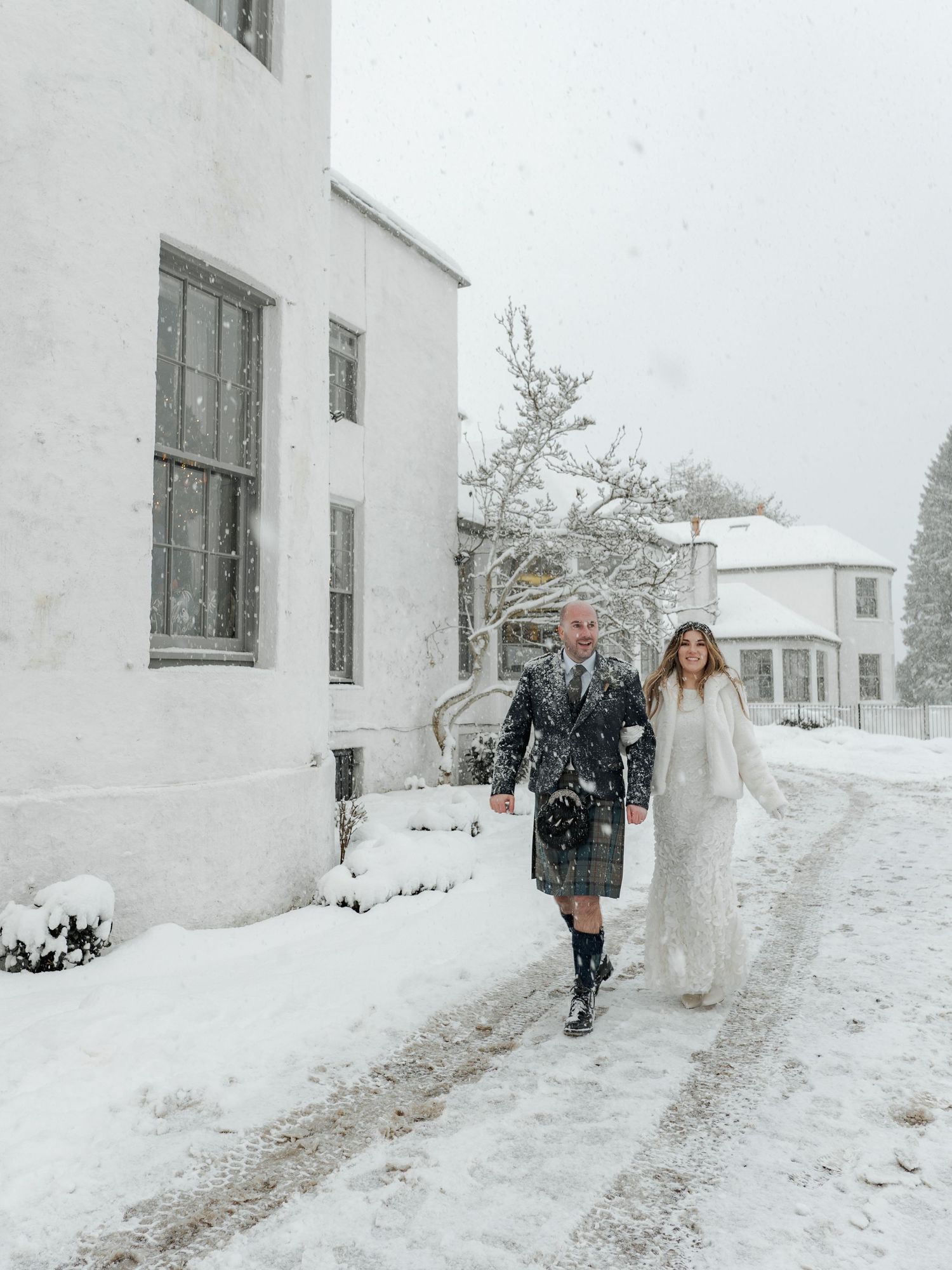 Bride and groom walking in heavy snowfall, groom wearing tartan kilt outside a snow-covered estate.