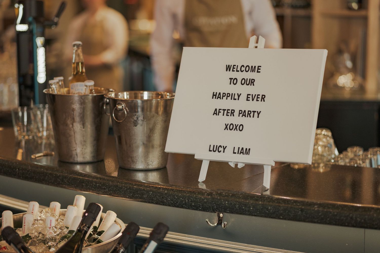 A welcome sign for an after party event sits on a bar counter next to ice buckets and glassware.