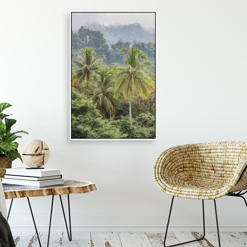 Framed tropical palm tree jungle print hanging on white wall above a rattan chair and wooden side table.