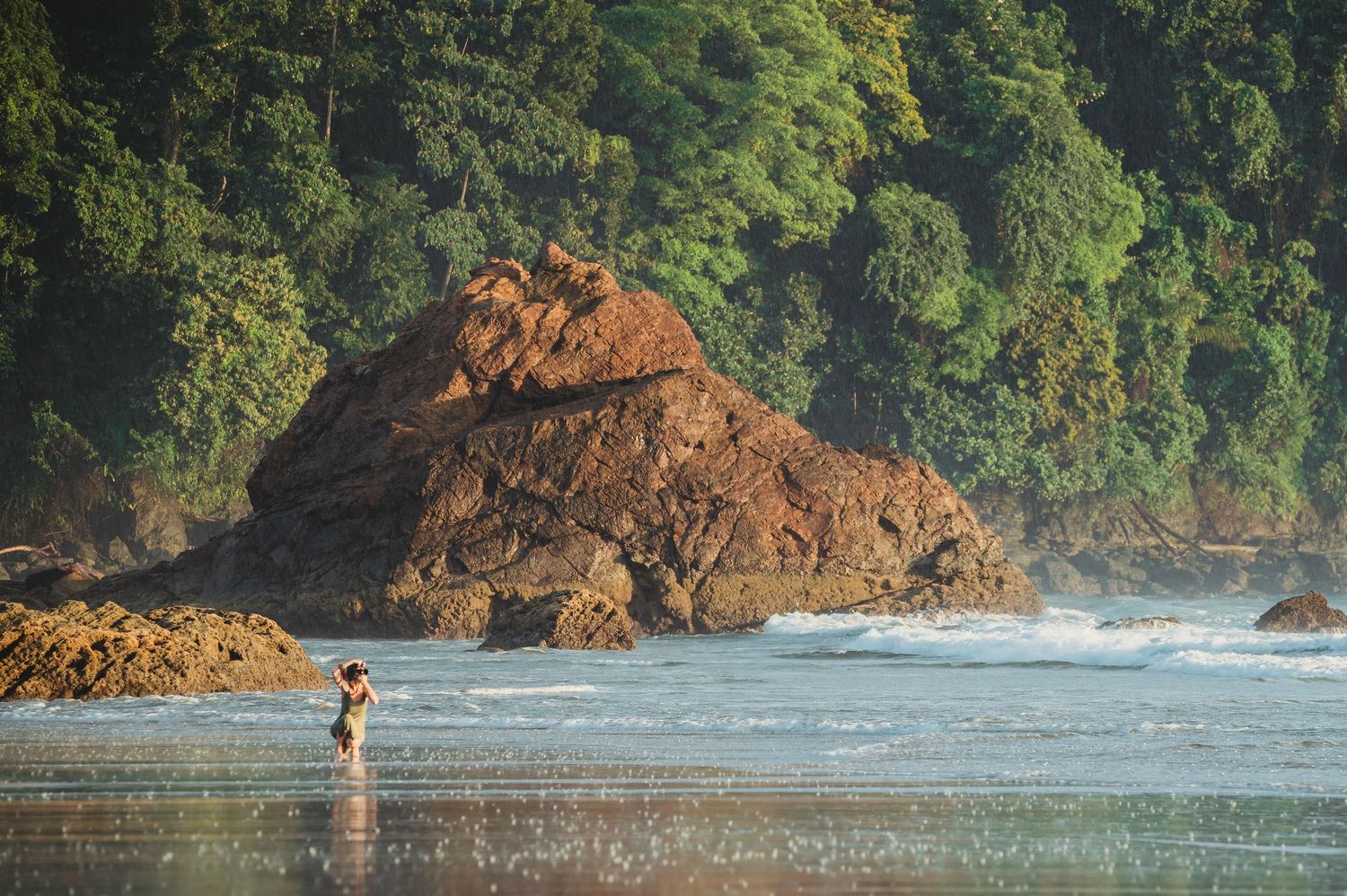 A lone person wades in shallow ocean waters surrounded by dramatic red rocks and lush tropical jungle coastline.