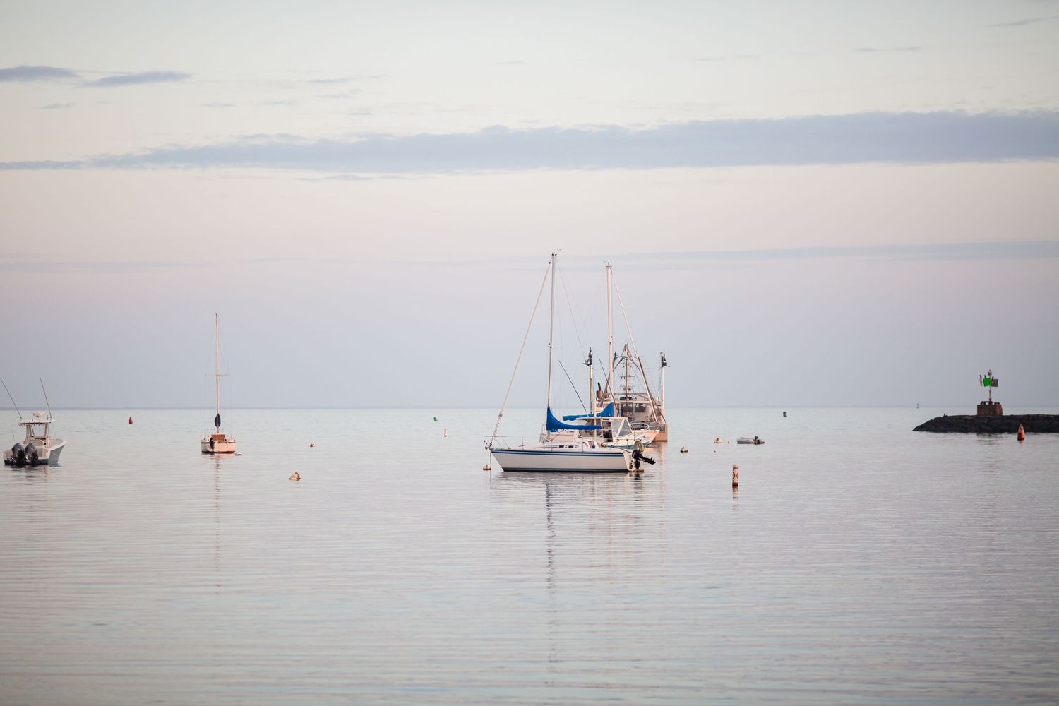 Sailboats anchored in a tranquil harbor during sunset with a lighthouse in the distance.