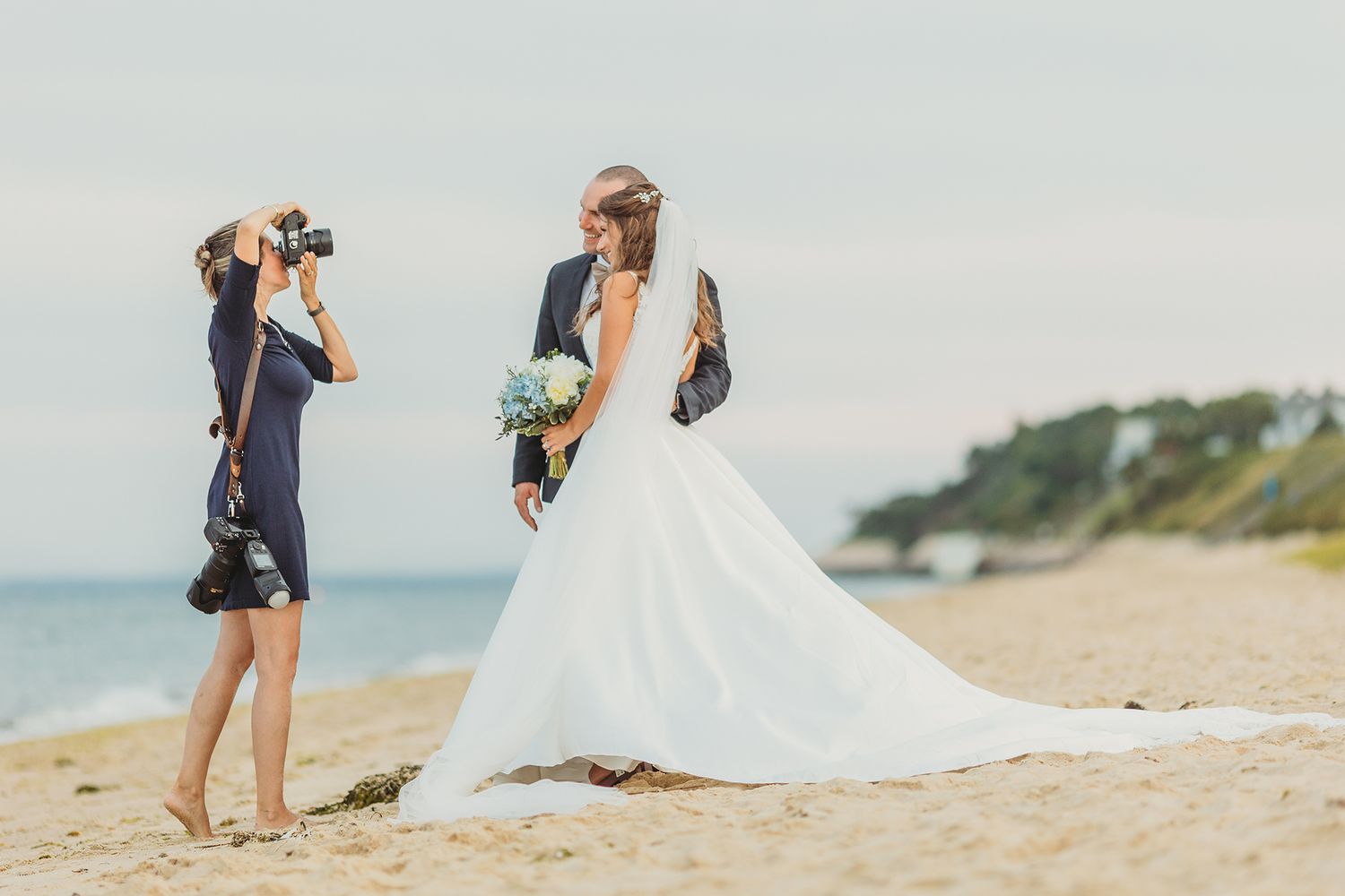 A photographer captures photos of a bride in a white dress on a sunny beach.