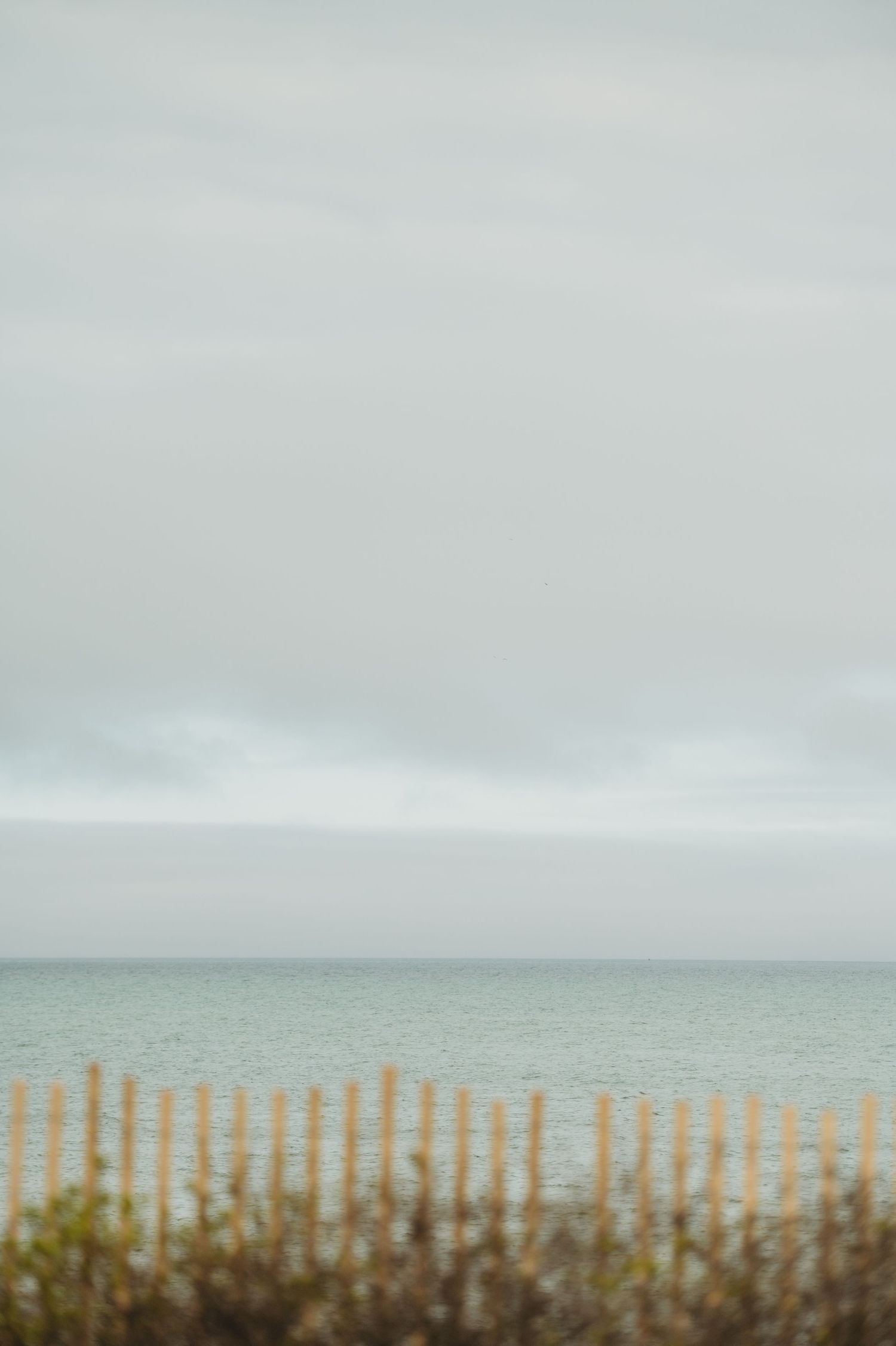 A wooden fence overlooks a calm ocean under an overcast sky on a quiet beach day.