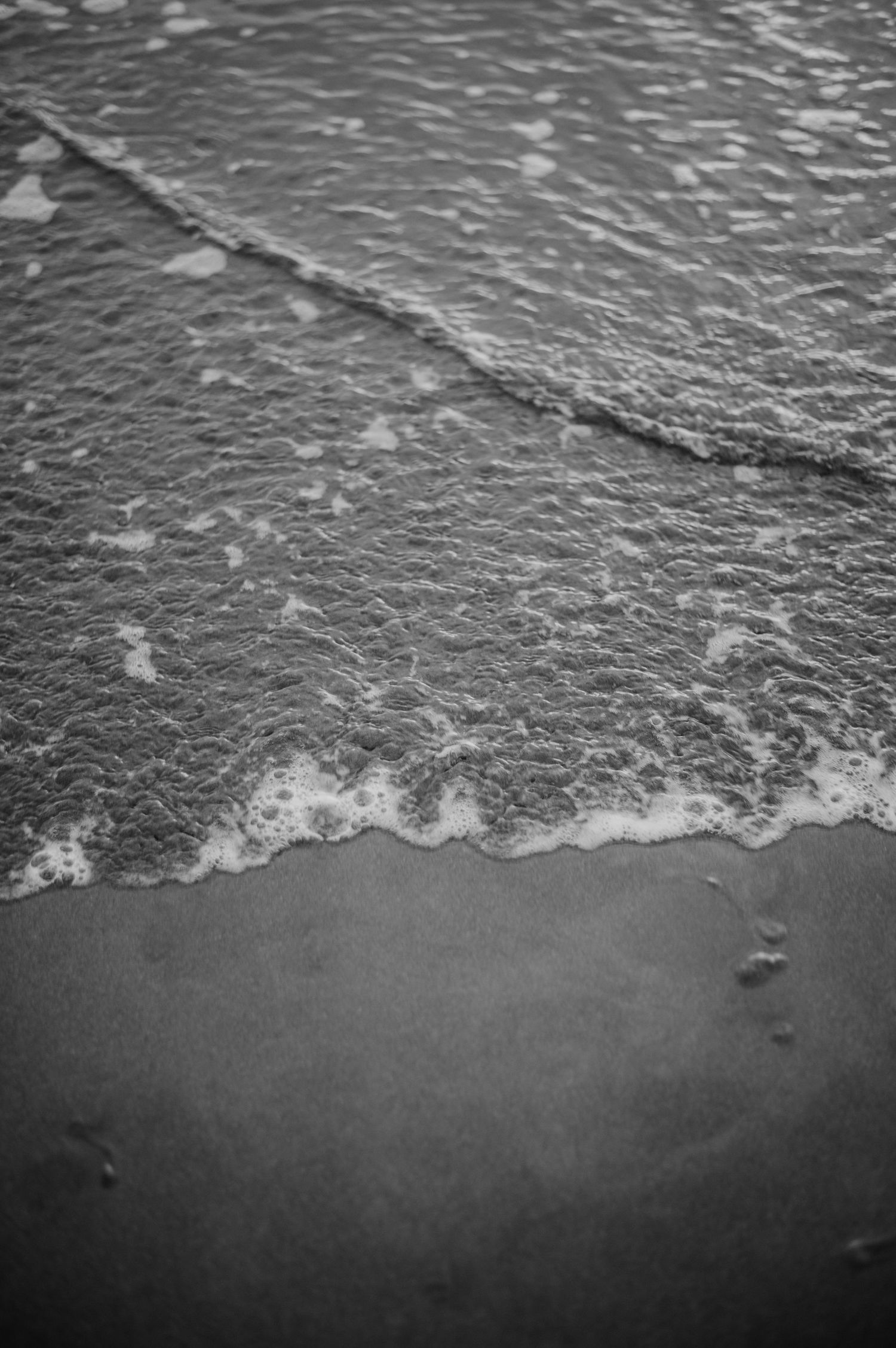 Black and white close-up of gentle waves washing onto a sandy beach.