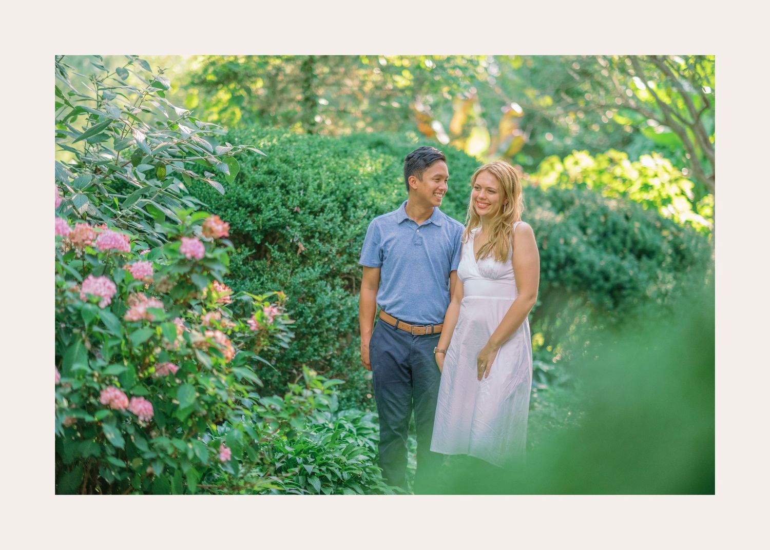 Couple standing together in a lush summer garden with blooming pink roses and green foliage.