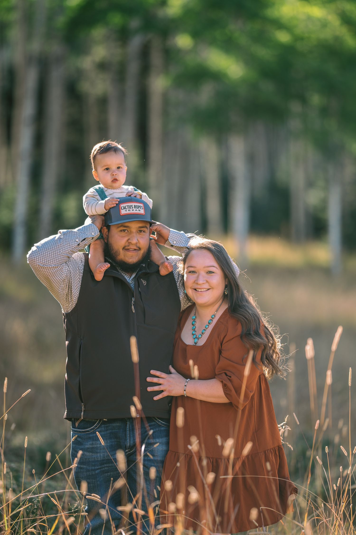A family of three poses together in tall grass with a forest backdrop.