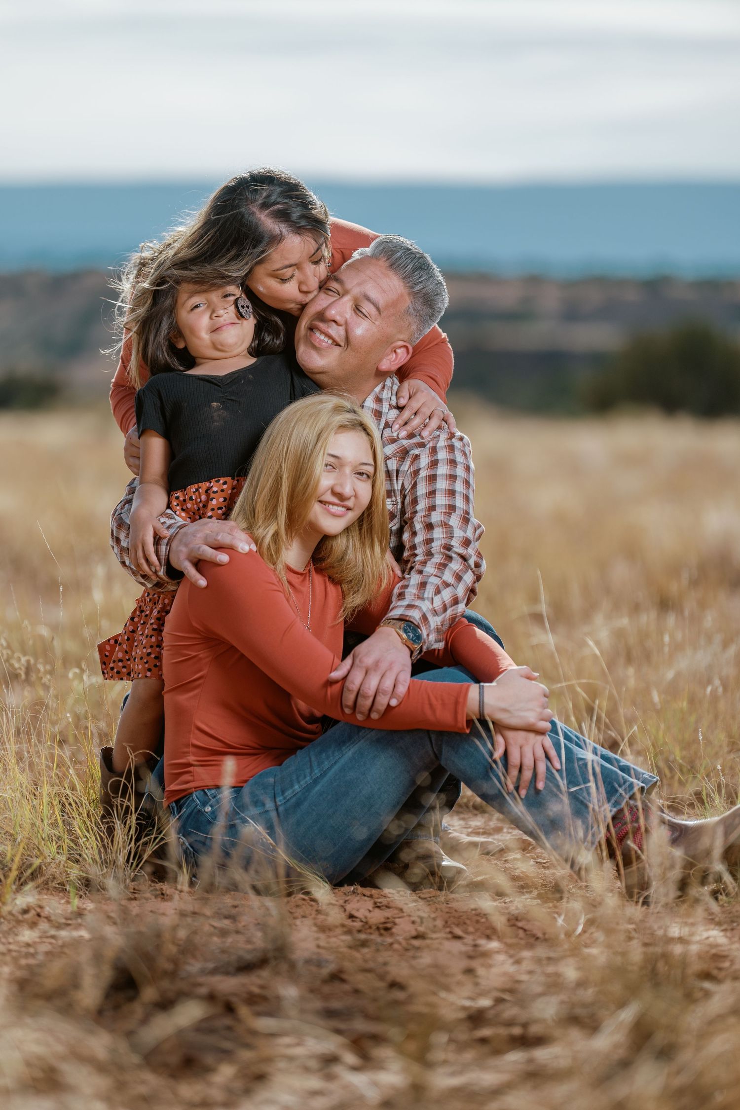 A family embraces while sitting in a golden field during sunset with distant mountains in the background.