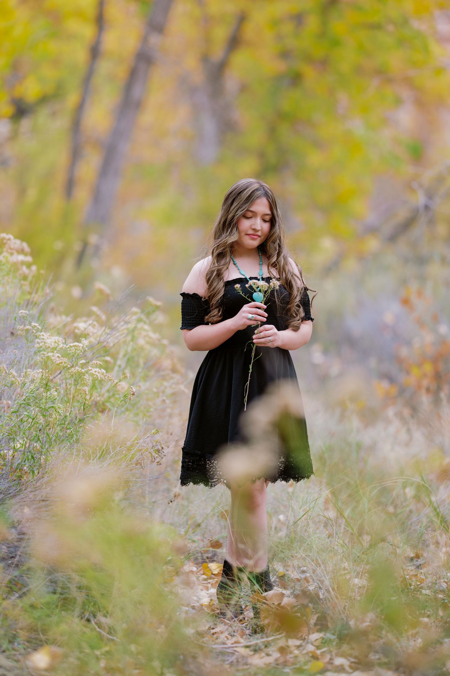 A person in a black dress stands in a dreamy autumn field holding flowers amid blurred golden foliage.