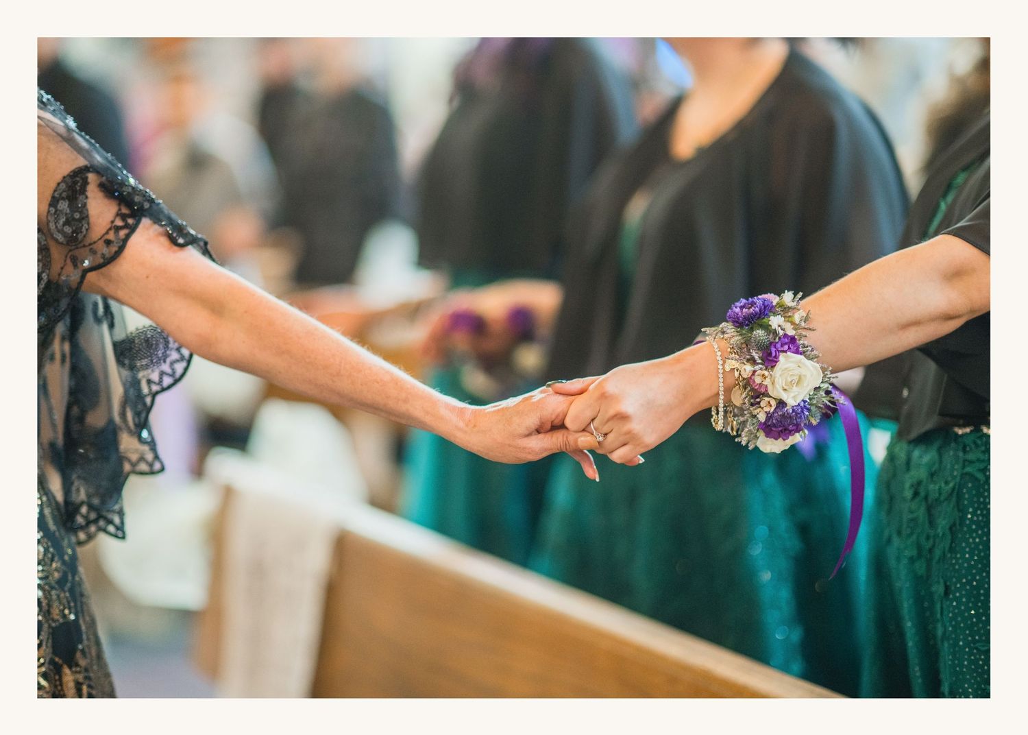 Two women holding hands at a wedding, one wearing a floral wrist corsage with purple flowers and teal ribbon.