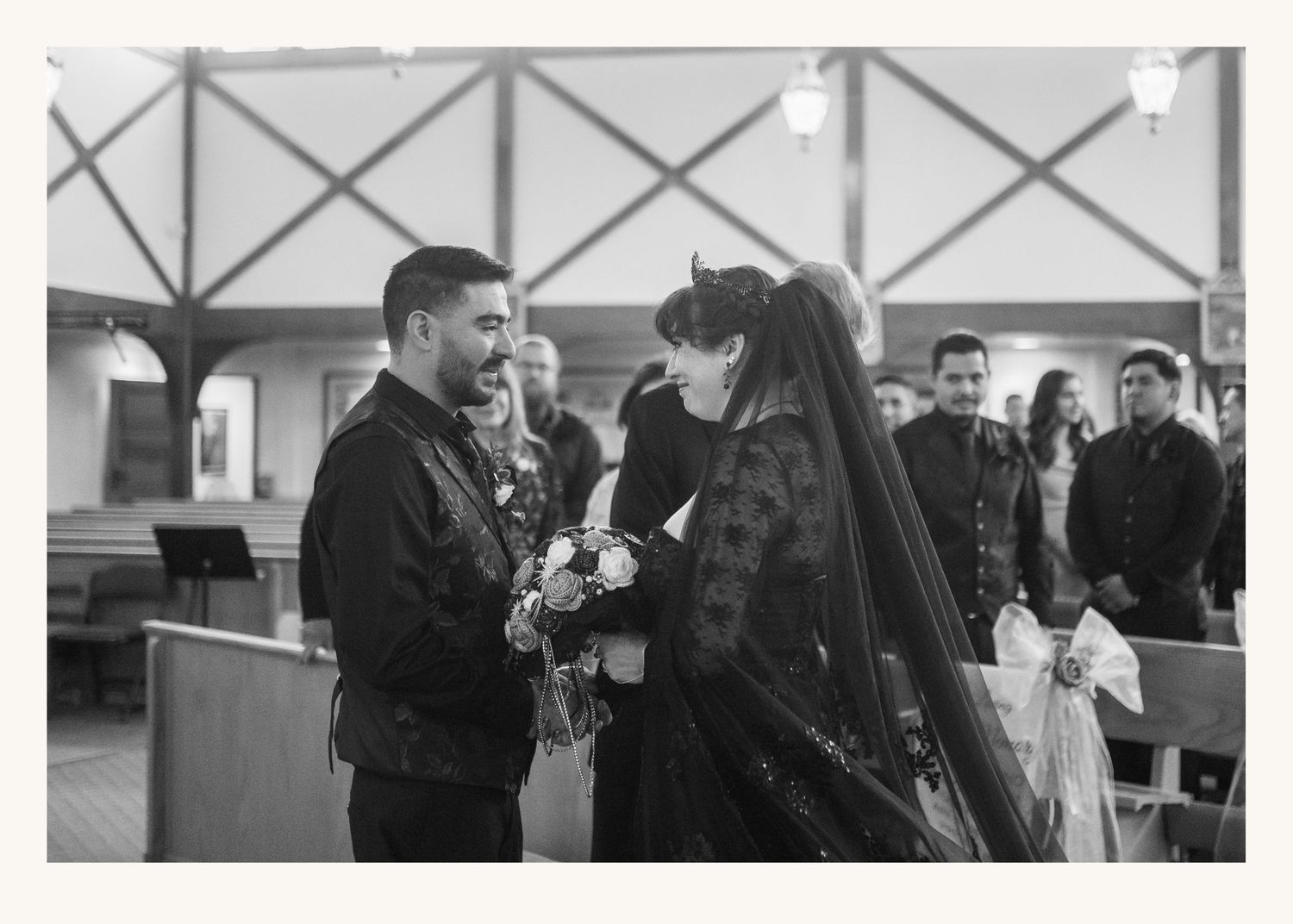 Black and white photo of a groom and gothic bride exchanging vows in a church aisle during their wedding ceremony.