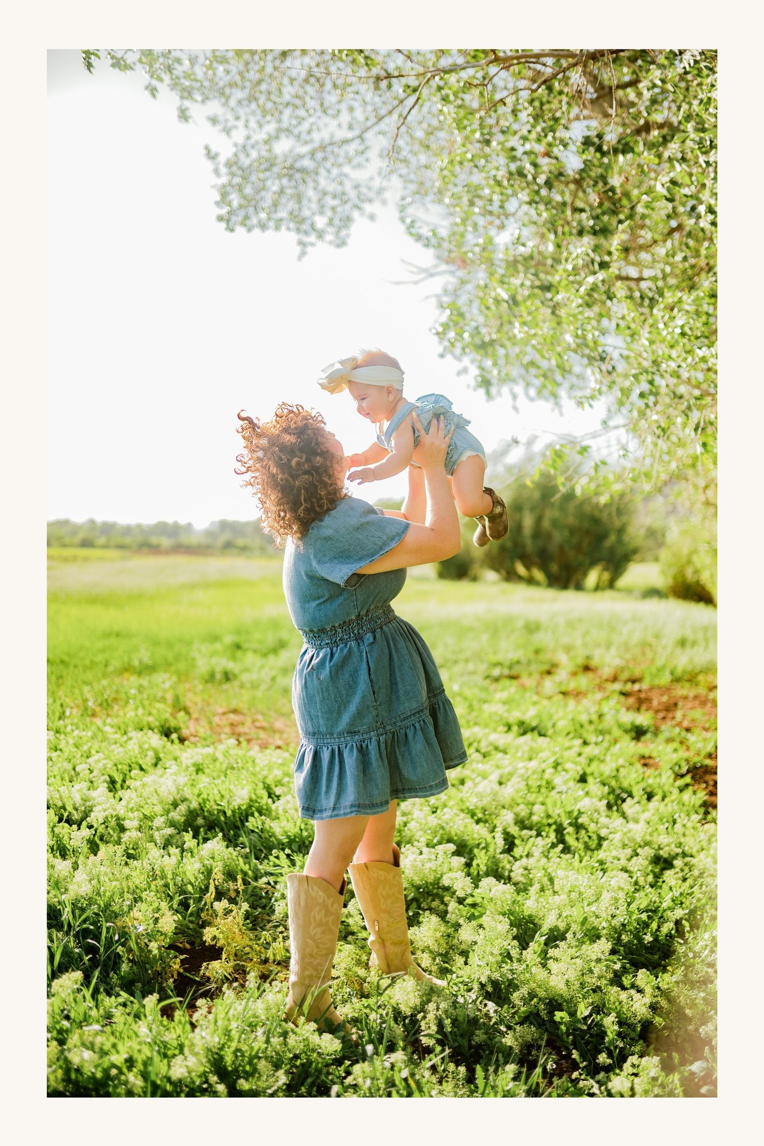 A curly-haired woman in a denim dress lifts a baby joyfully in a sunny green field.