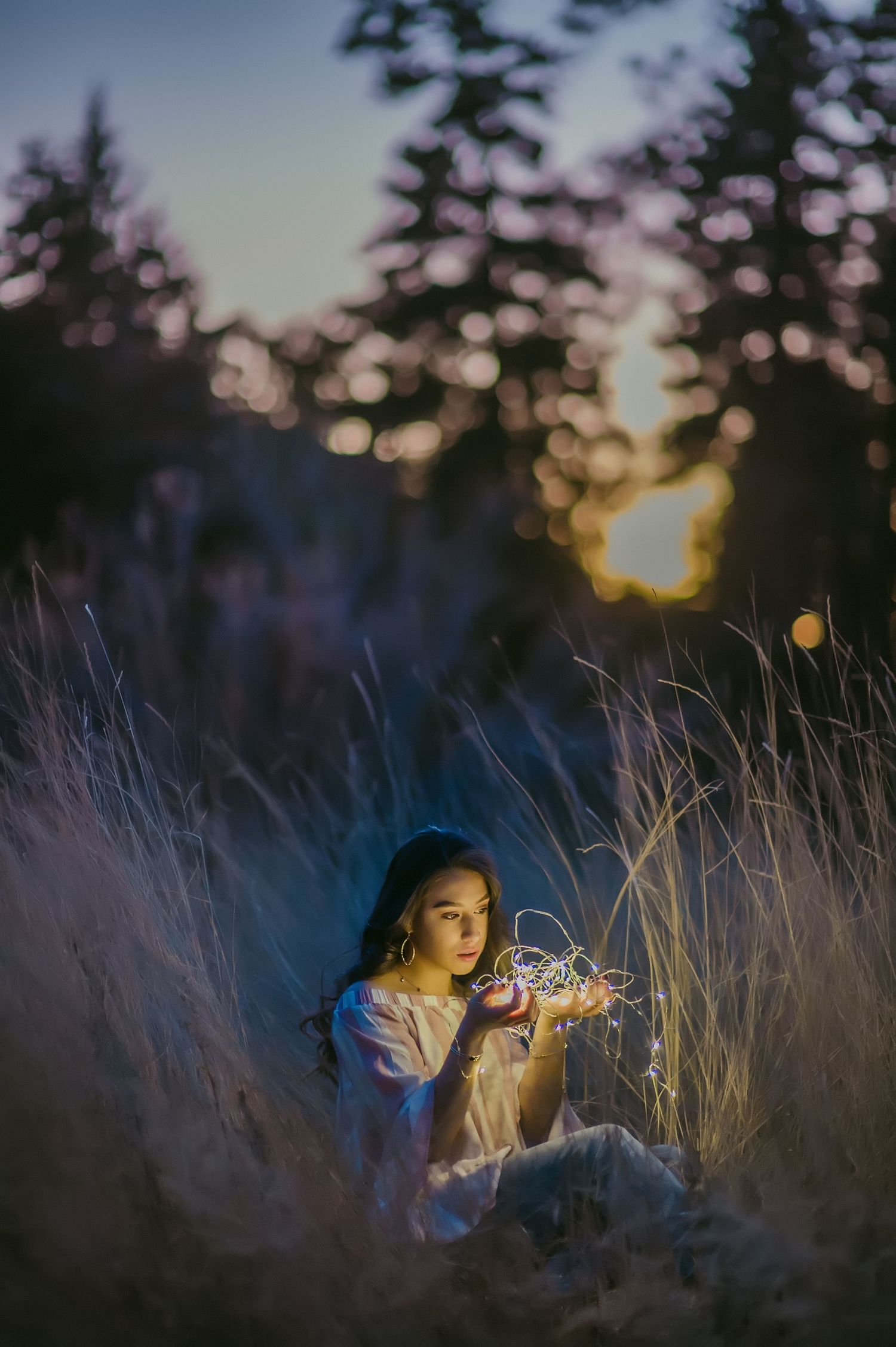 A young woman sits in tall grass at dusk, holding glowing fairy lights in her hands, with bokeh trees in the background.