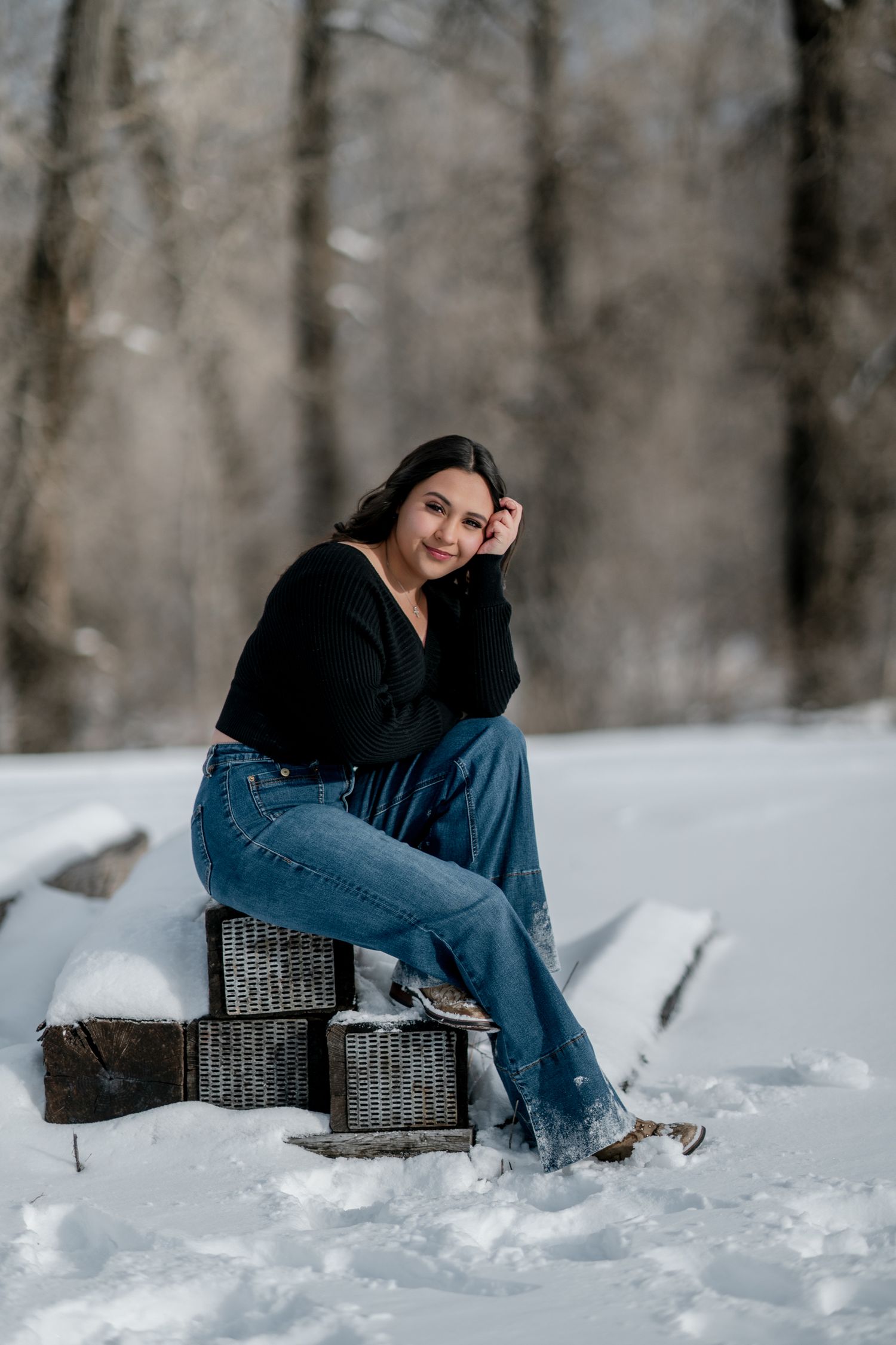 A person in blue jeans and dark top sits casually on a wicker chair in snowy winter landscape.