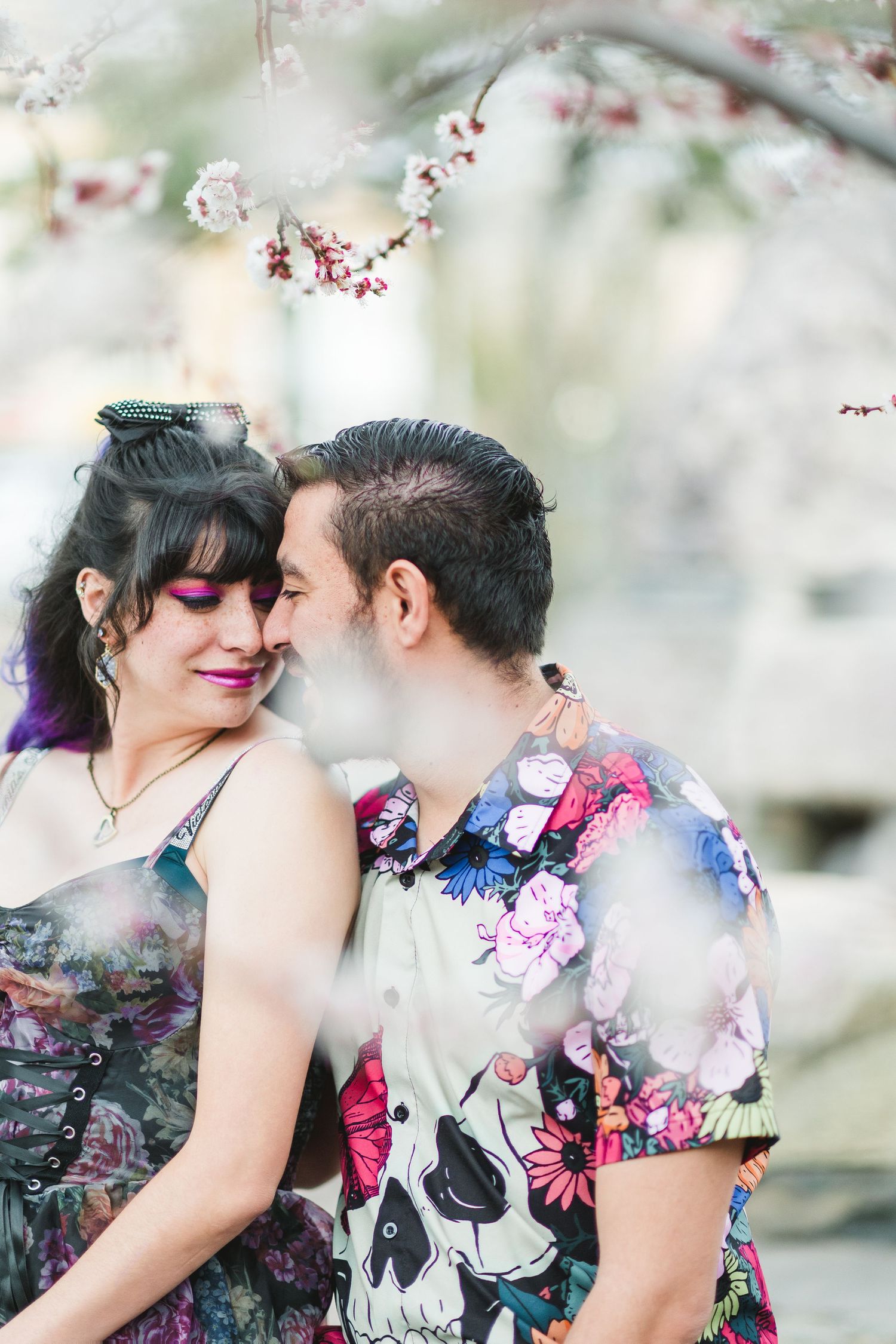 Couple sharing a romantic moment under cherry blossom tree, wearing colorful floral and skull print outfits.