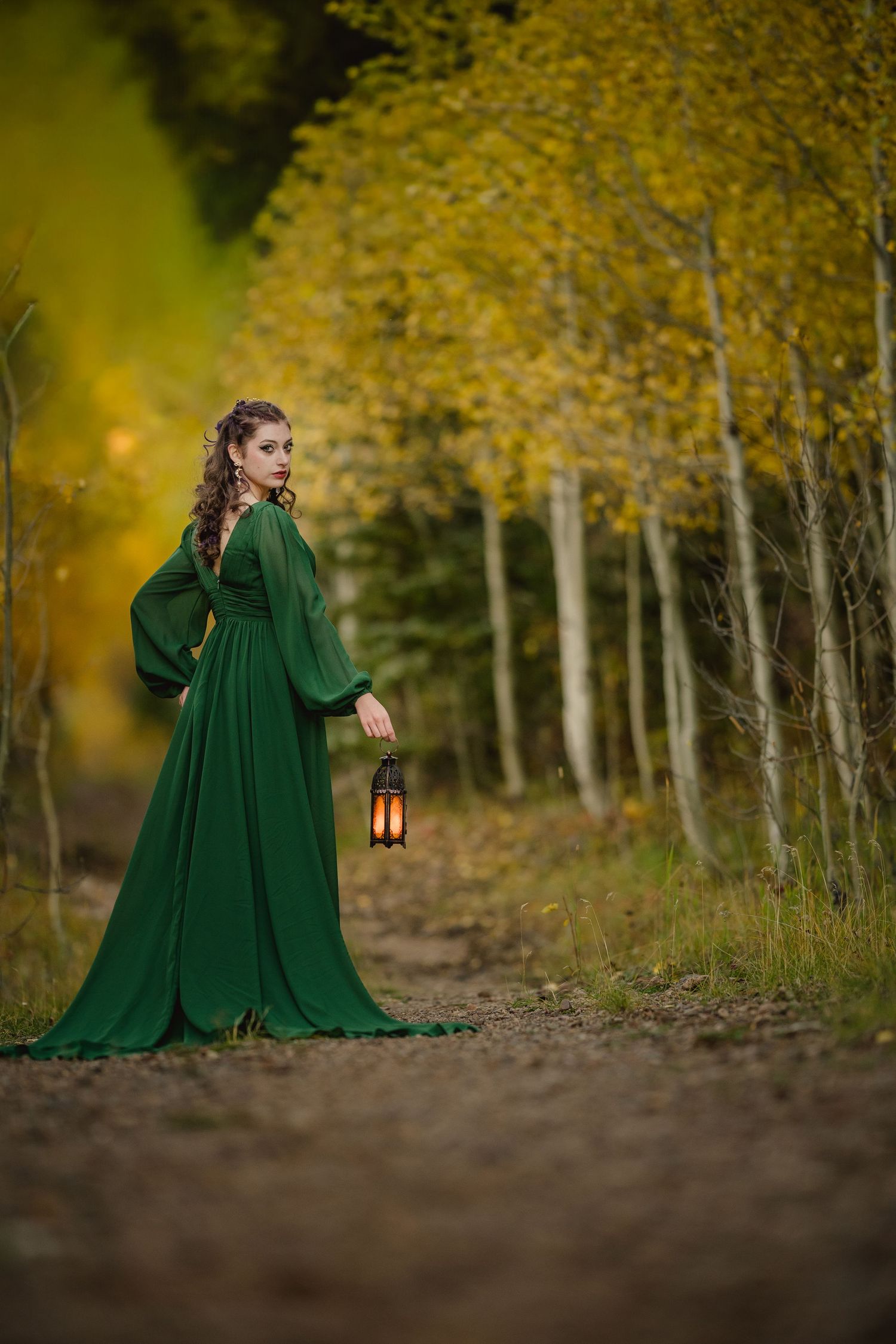 A figure in an emerald green cloak holds a lantern while walking through a golden autumn forest setting.