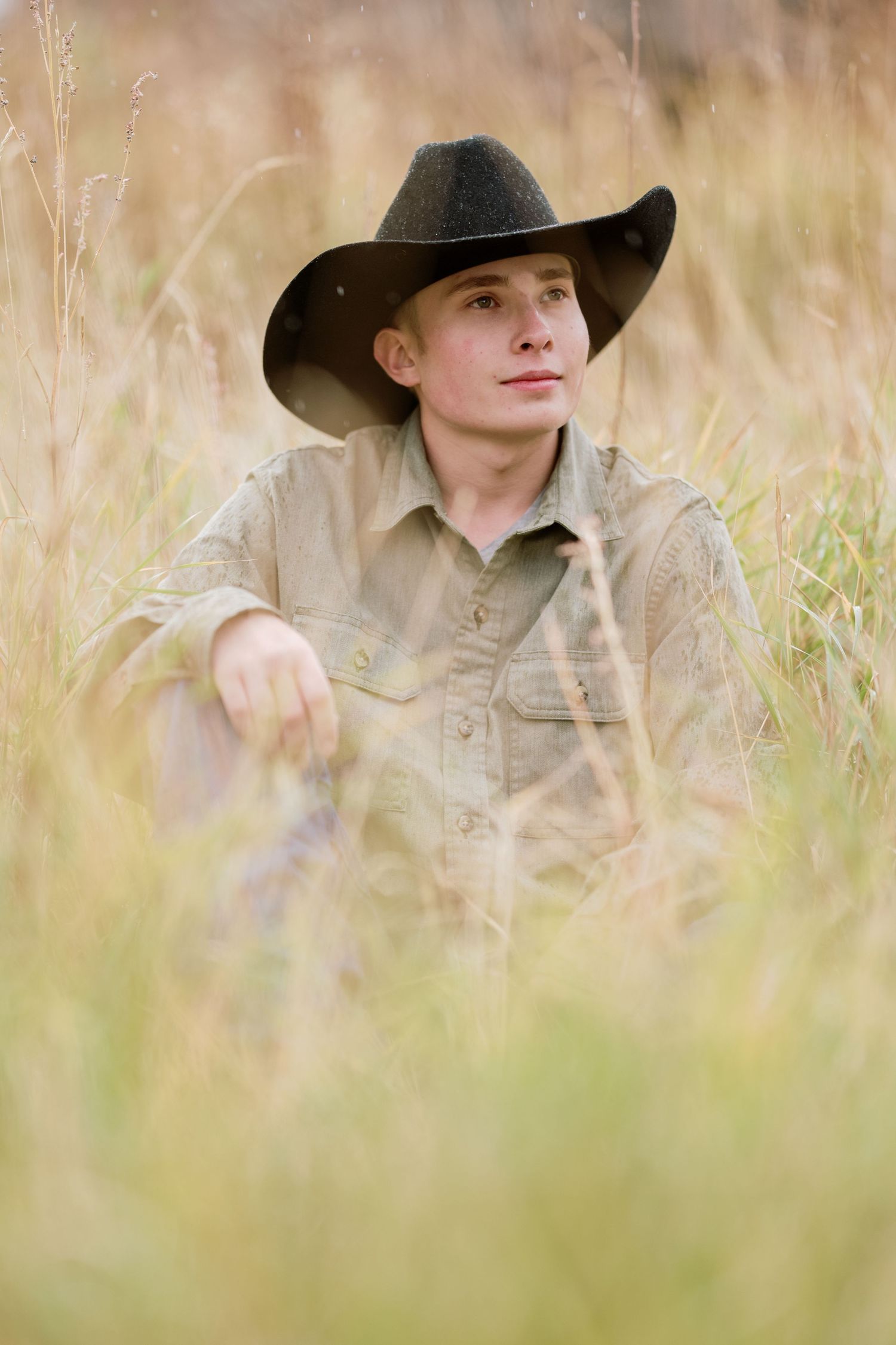 A contemplative young cowboy in a black hat sits in tall prairie grass wearing a tan shirt.