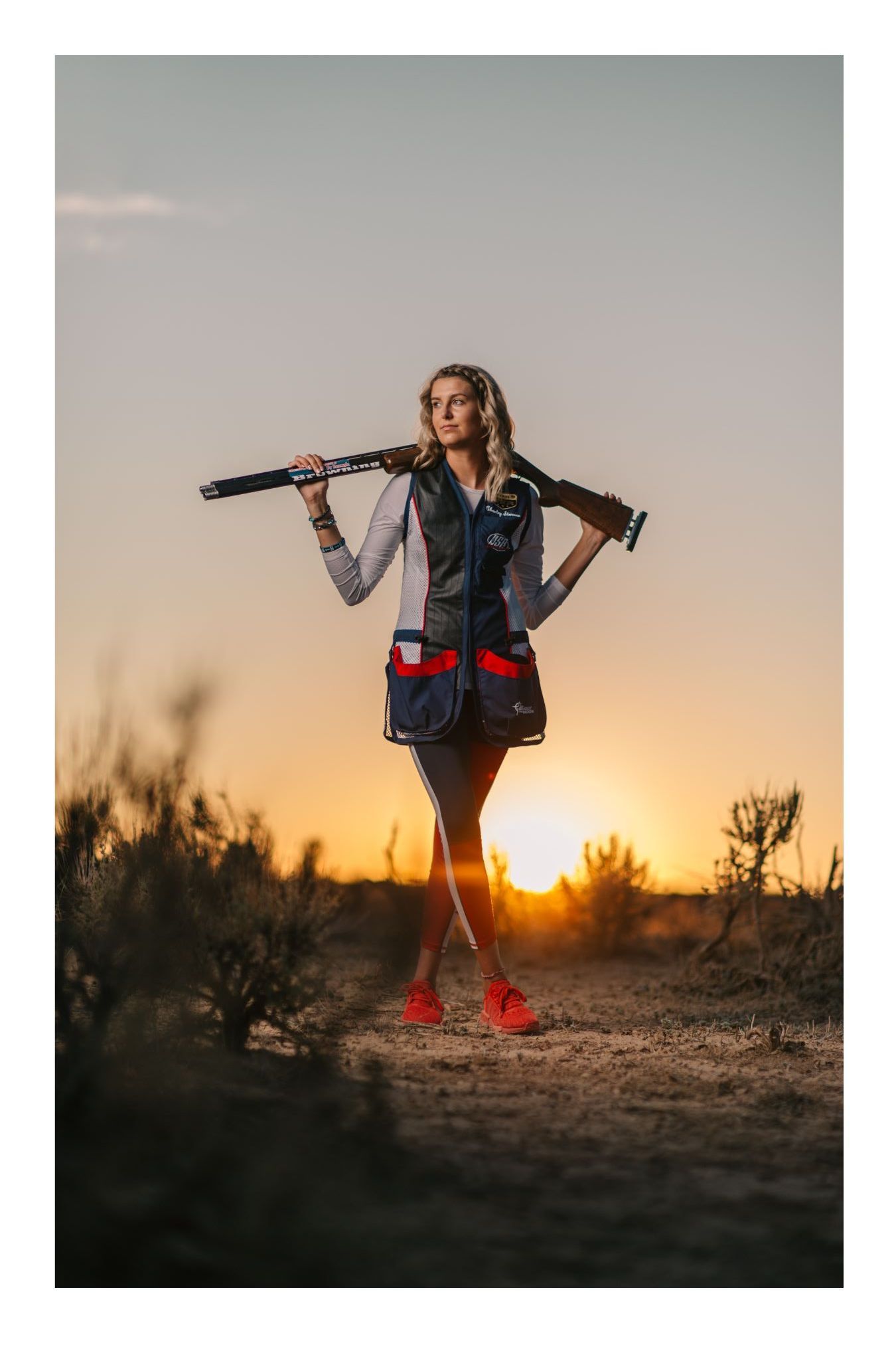 Woman holding a shotgun over her shoulder in a shooting vest at sunset in a desert landscape.