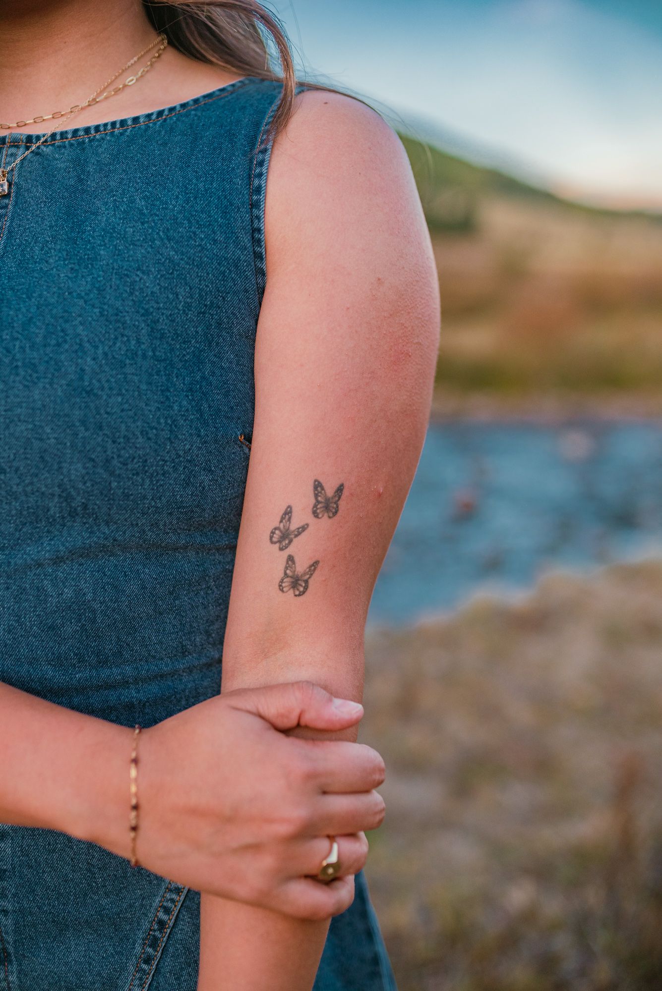 Woman in denim dress with three small butterfly tattoos on her forearm, standing near a scenic outdoor backdrop.