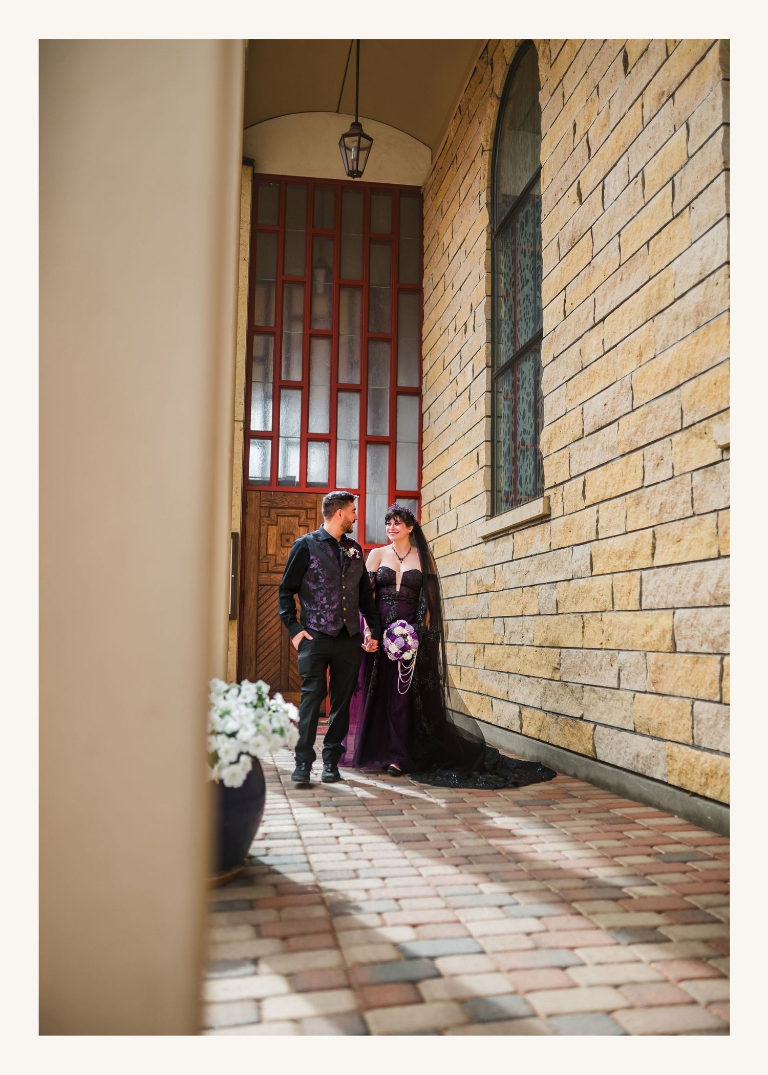 Couple in elegant gothic wedding attire posing by a brick building entrance with a ornate wooden door.
