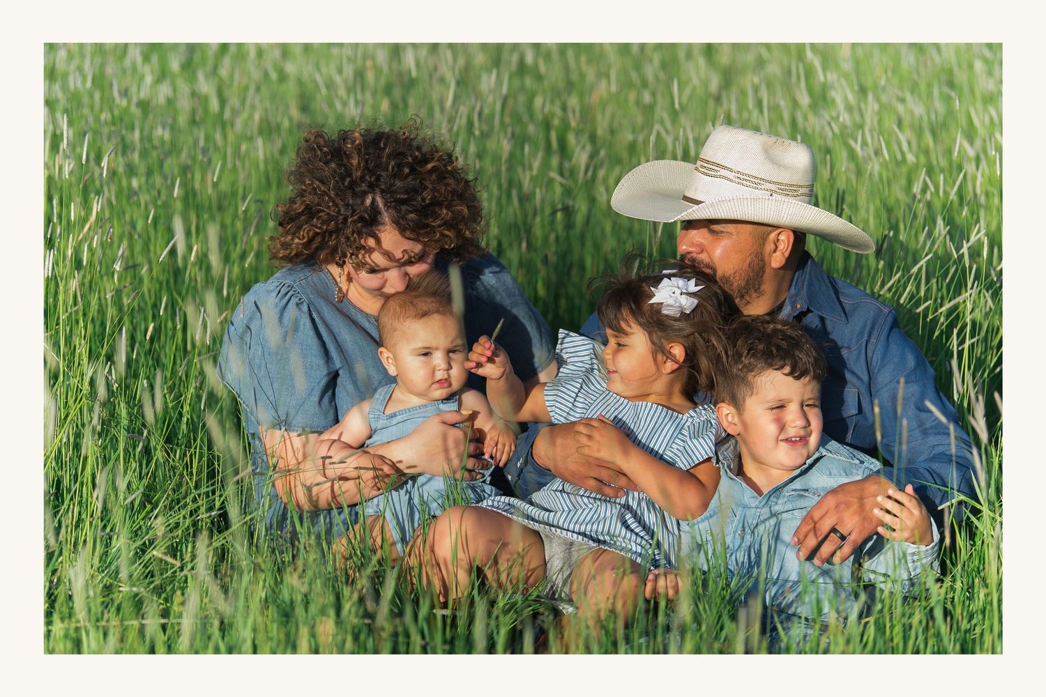 Family of five sitting in tall green grass field, parents holding children, wearing denim and cowboy hat.