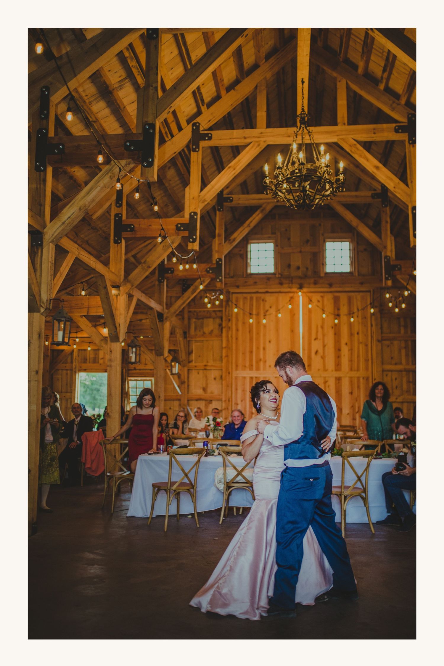 Couple shares first dance in rustic barn wedding venue with string lights and chandelier overhead.