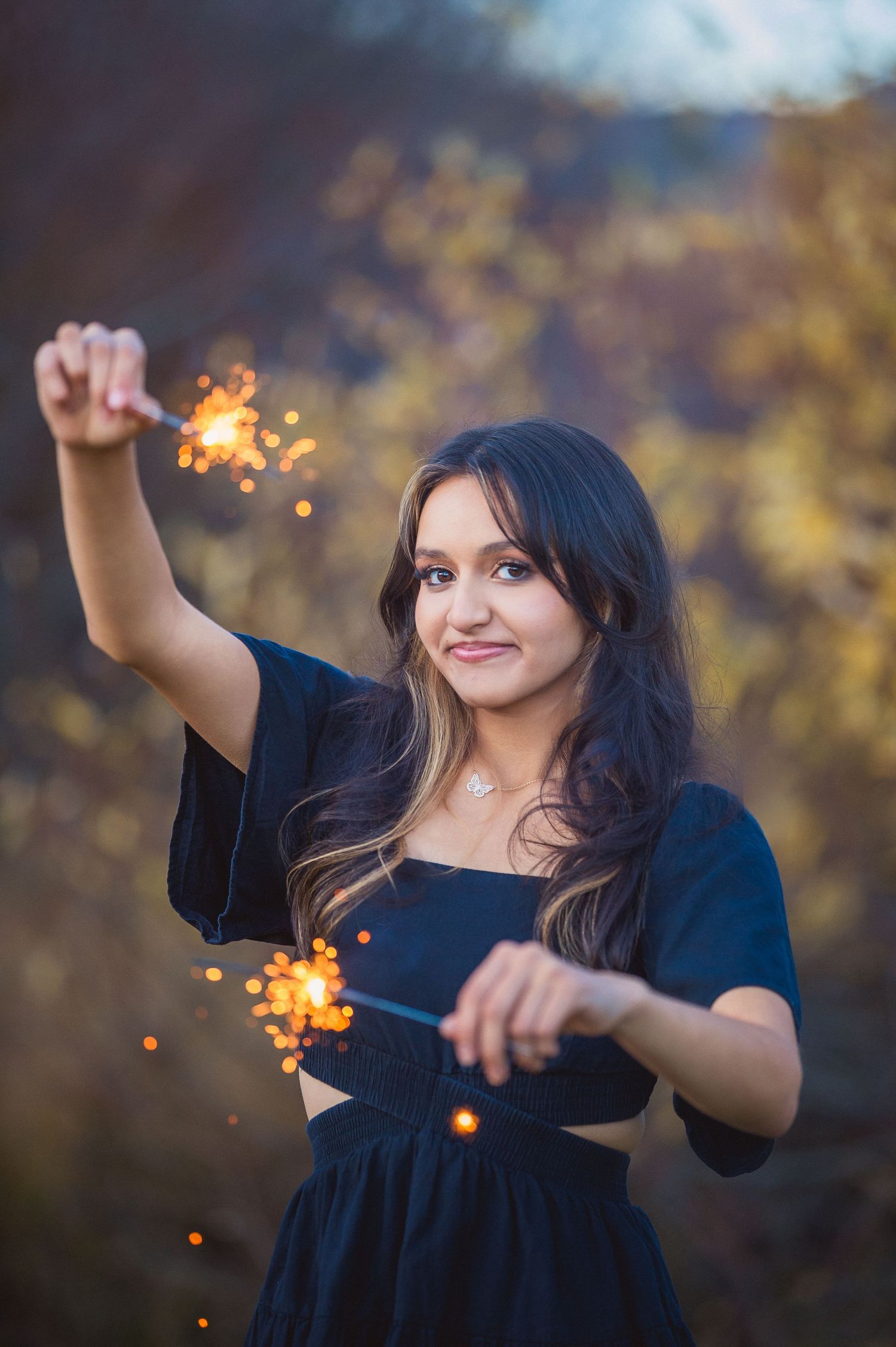 A person in a dark dress waves sparklers creating light trails against a blurred autumn background.