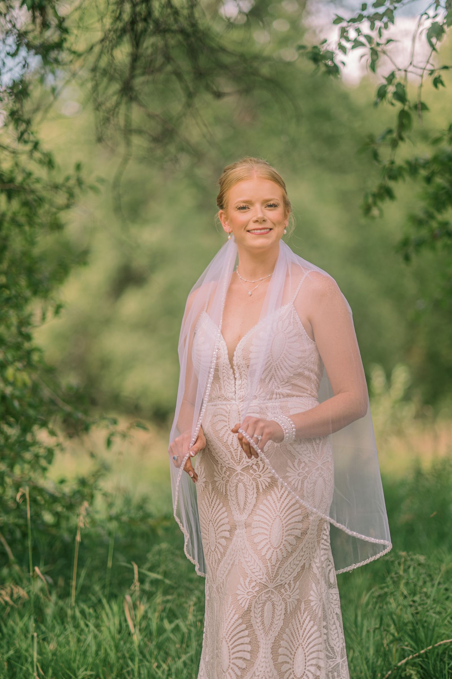 Bride in lace wedding gown and veil smiling outdoors surrounded by lush green trees.