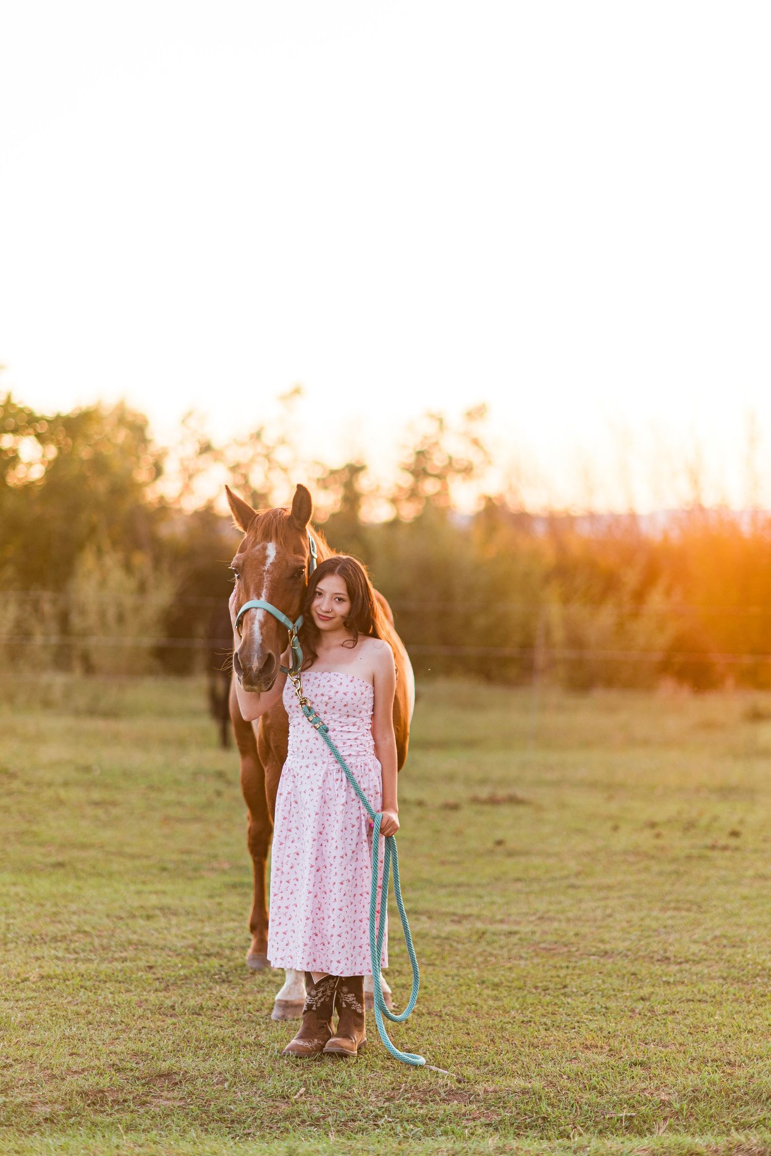 Person in a polka dot dress stands with a brown horse in a sunlit field at golden hour during sunset.