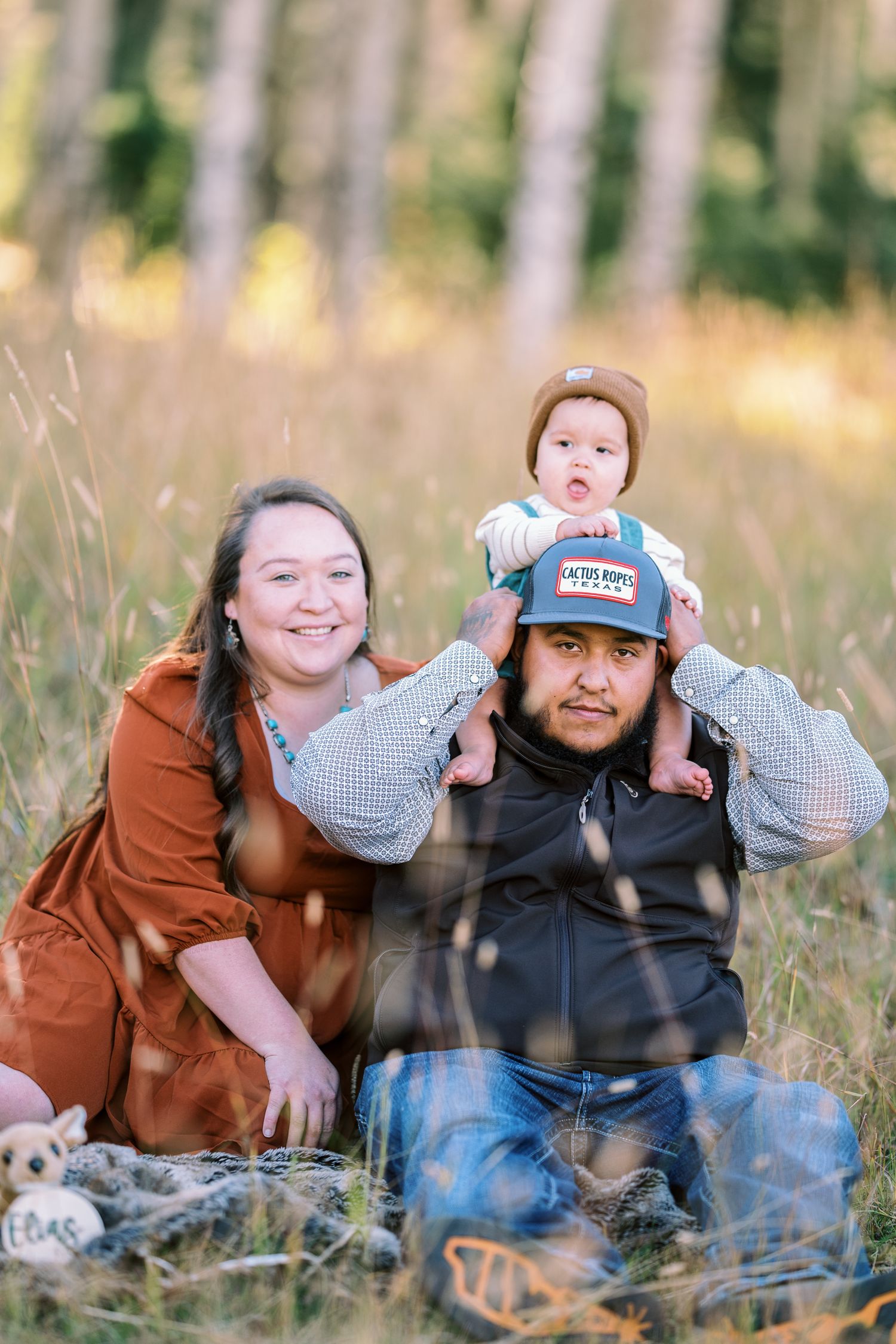 A family sits together on a blanket in a field with tall grass and trees.