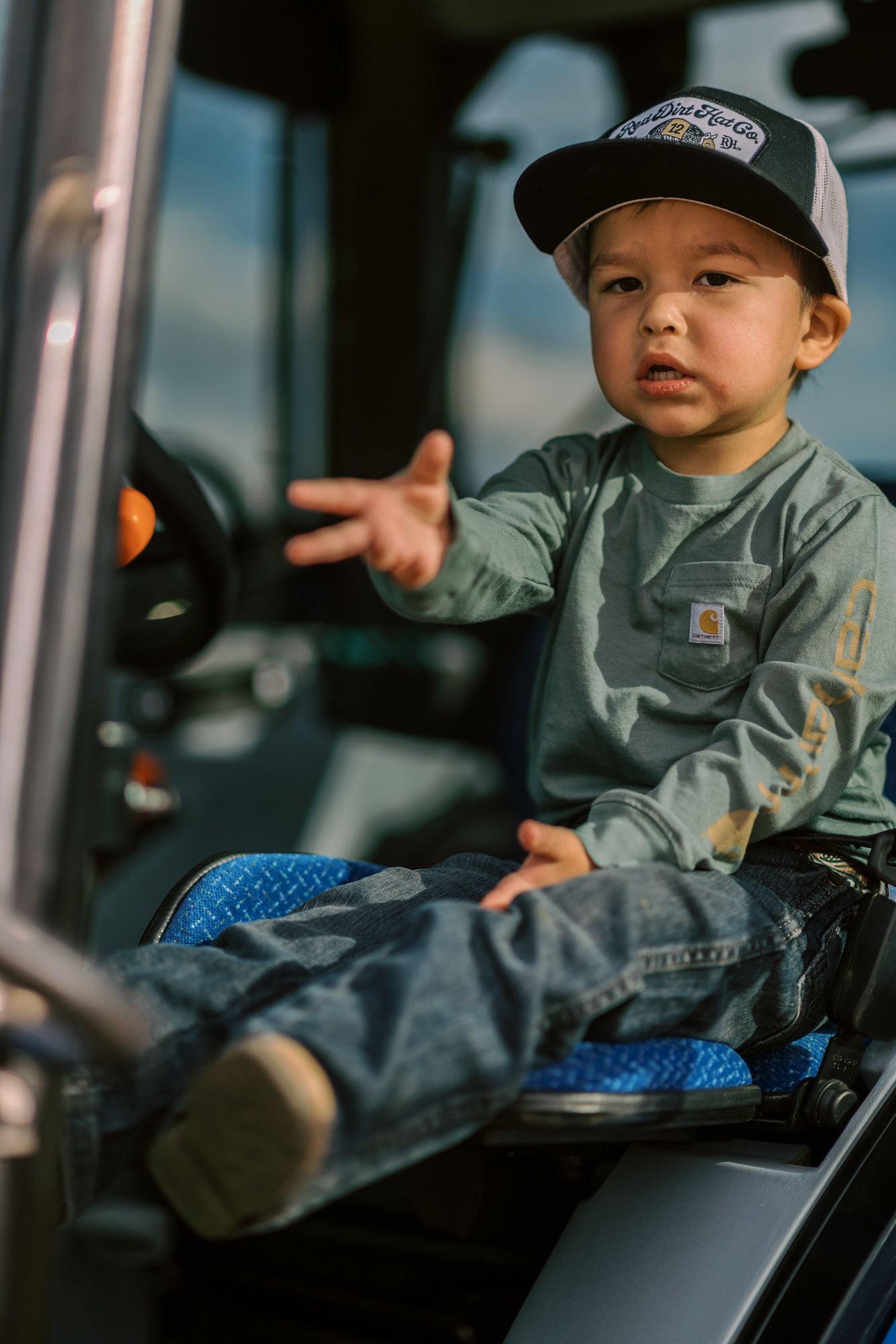 Young boy in Carhartt shirt and trucker hat sitting in tractor cab reaching toward steering wheel.