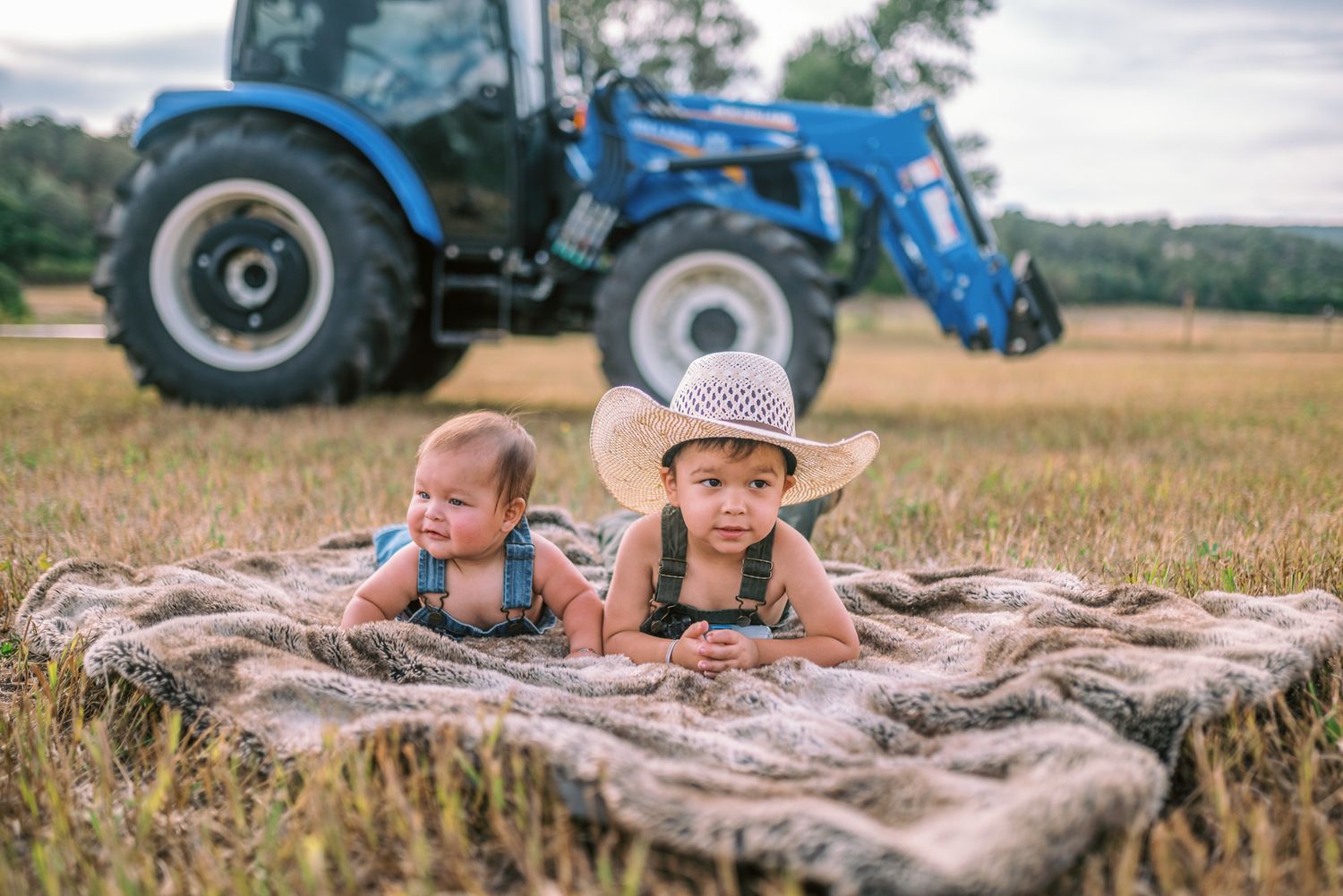 Two babies lying on a blanket in a farm field with a blue tractor in the background.