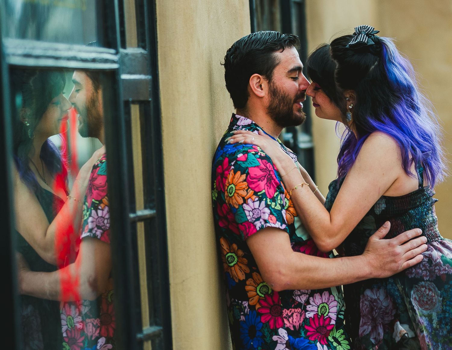 Couple sharing a romantic kiss outdoors, man in floral shirt embracing woman with purple hair near a glass window.