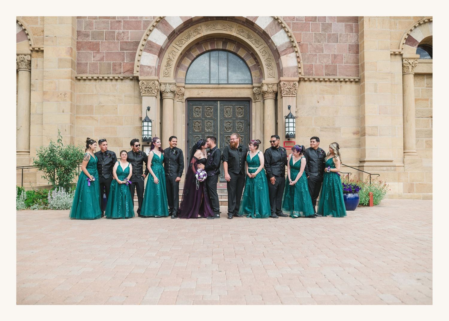 Wedding party in emerald green gowns and black suits posing before a historic stone church entrance.