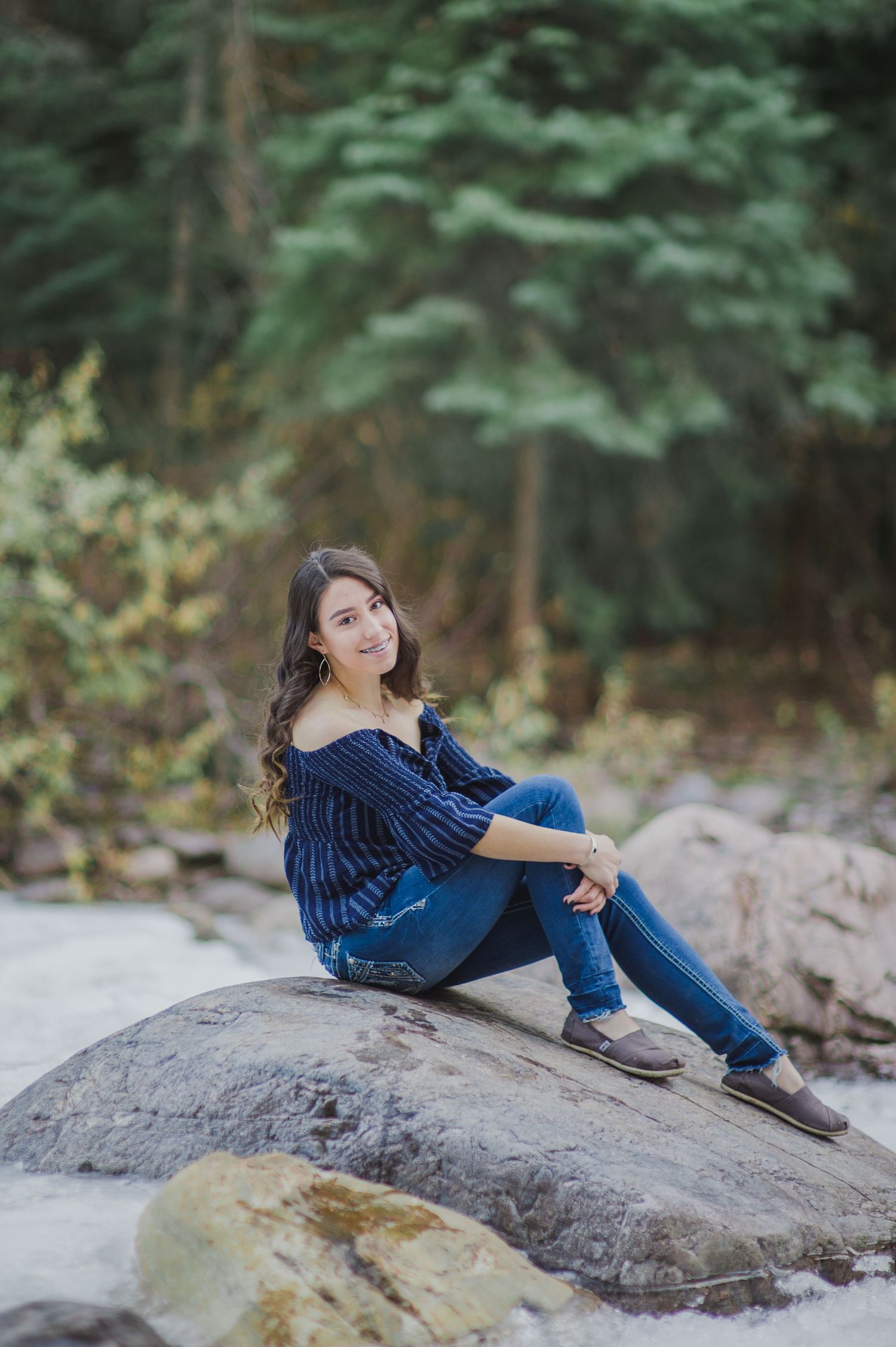 Young woman in blue off-shoulder top and jeans sitting on rocks outdoors with lush green trees in background.