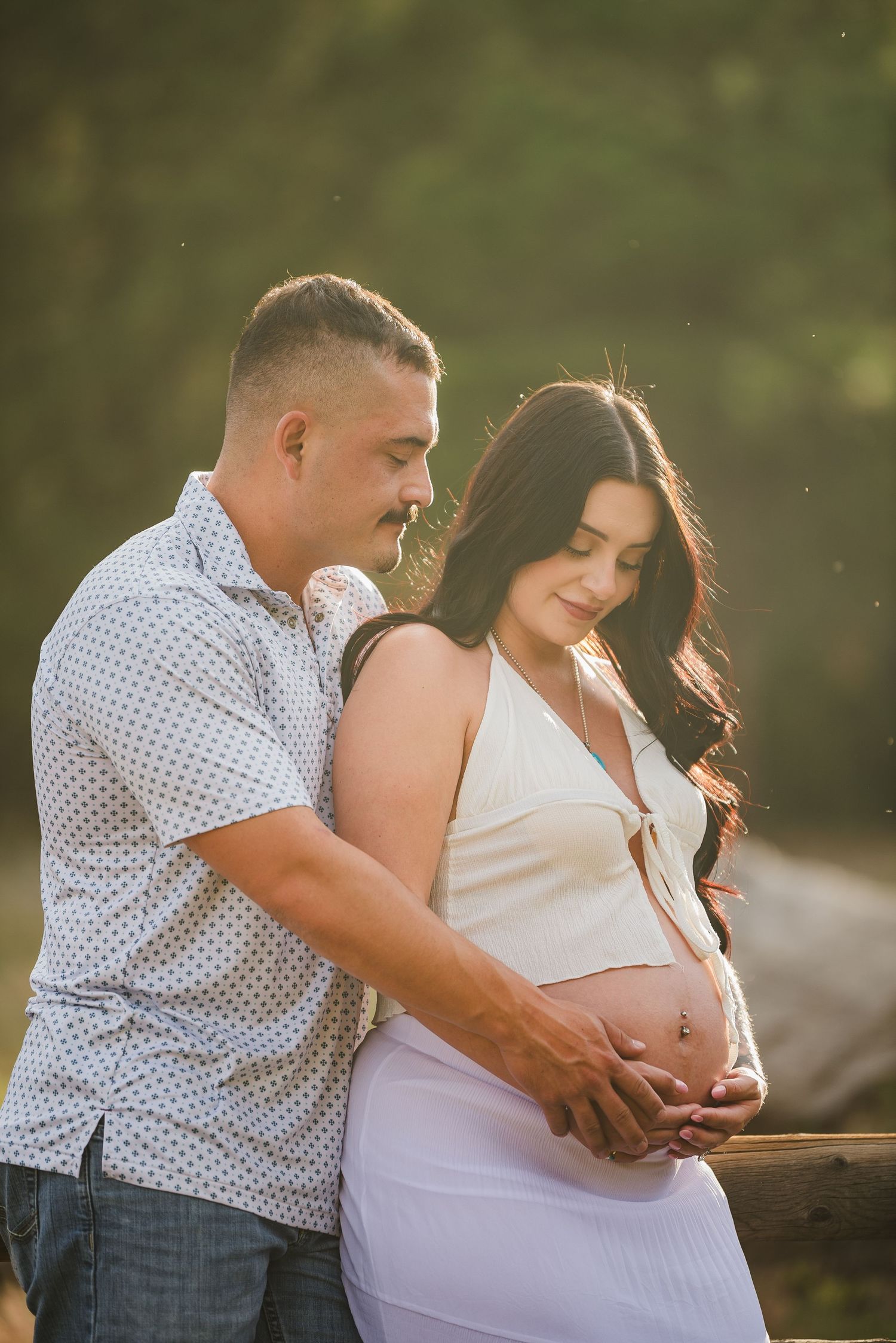 Couple tenderly cradling pregnant belly outdoors in golden sunset light, man embracing woman in white maternity outfit.