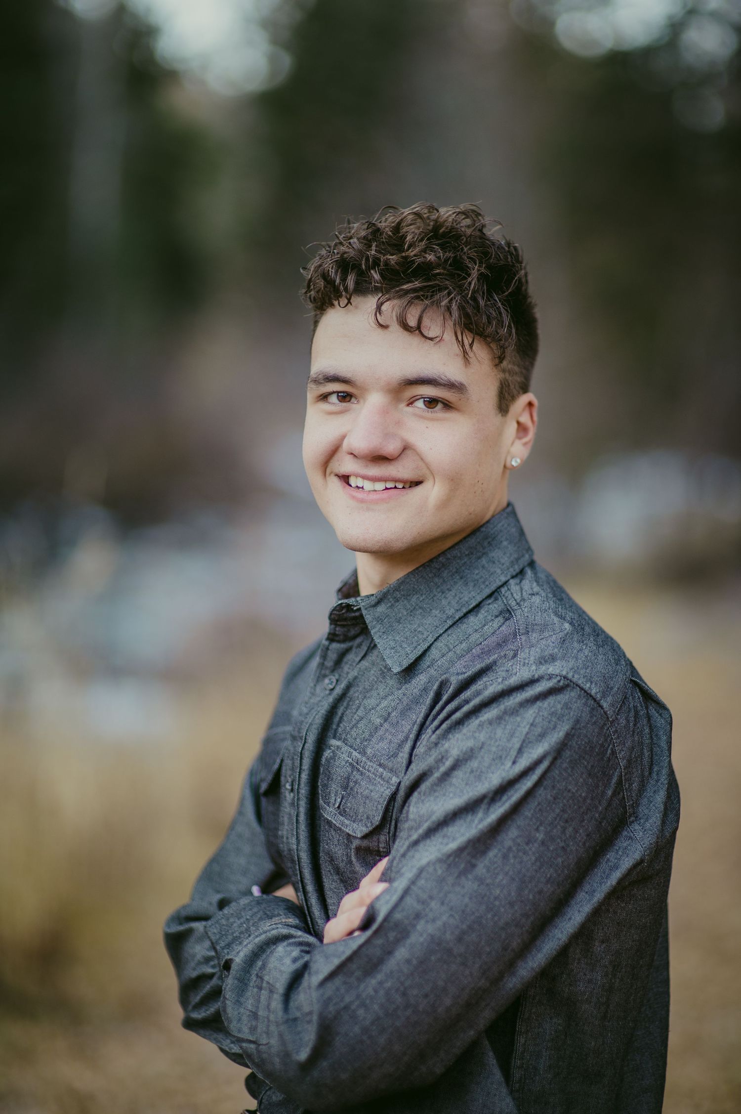 Young man with curly hair smiling outdoors, arms crossed, wearing a gray denim shirt.