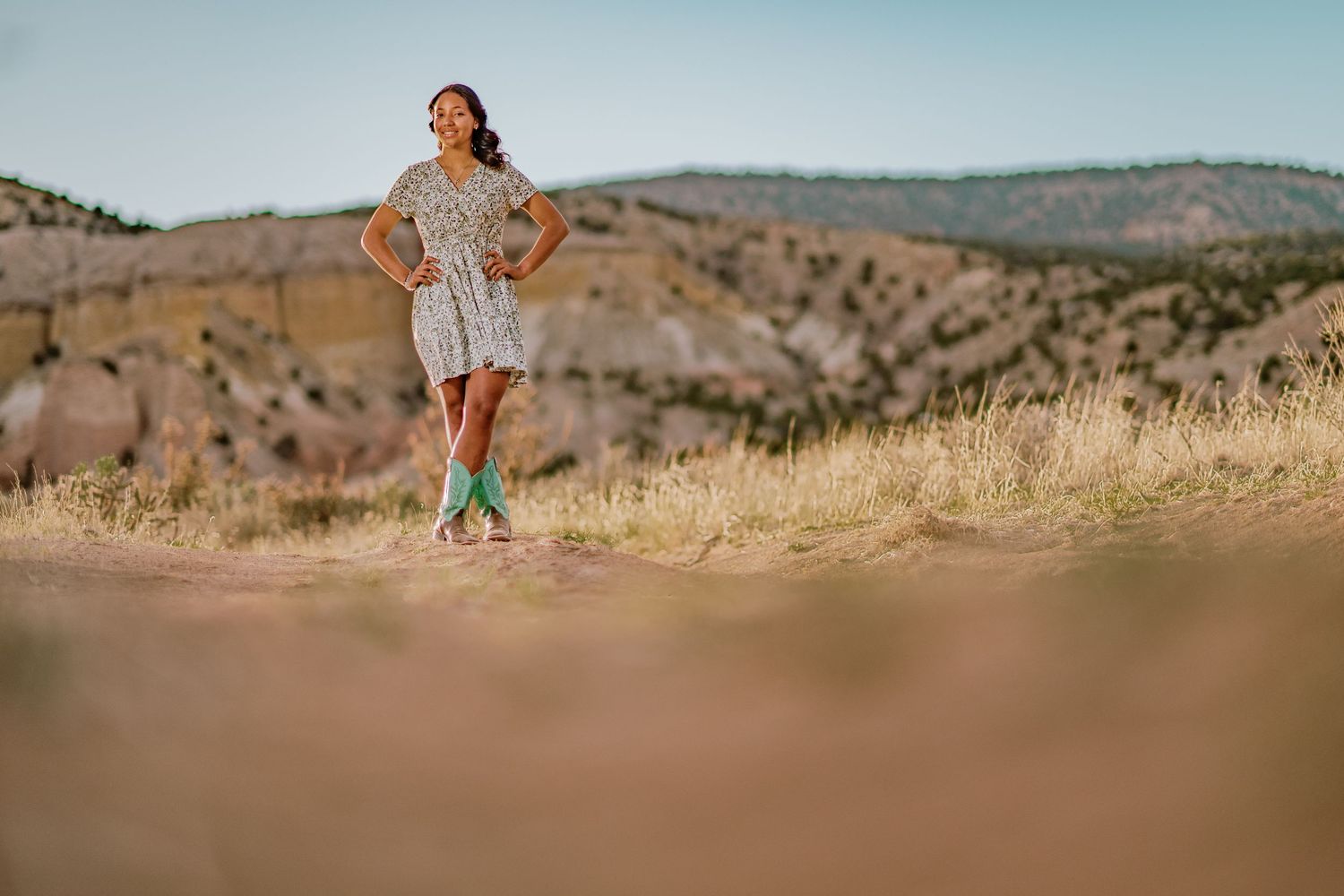 Runner in a flowy dress stands on a hilltop trail against a backdrop of rolling golden hills in warm sunlight.