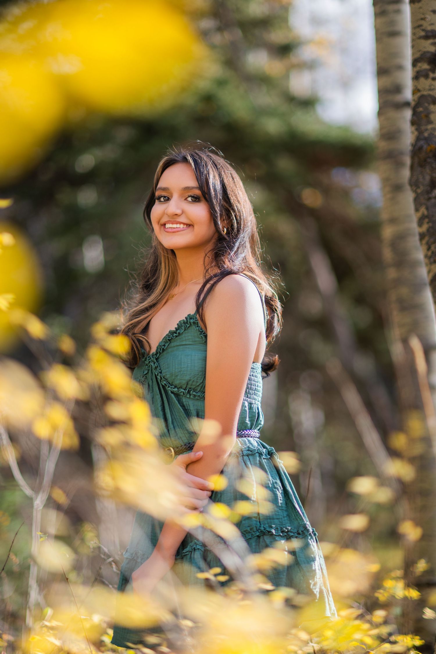 A woman in a green dress poses outdoors among yellow flowers and trees during a portrait photography session.