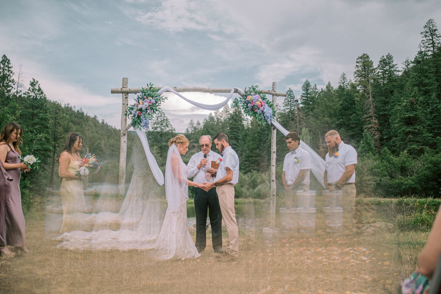 Outdoor wedding ceremony under floral arch with bride, groom, and officiant surrounded by lush forest backdrop.