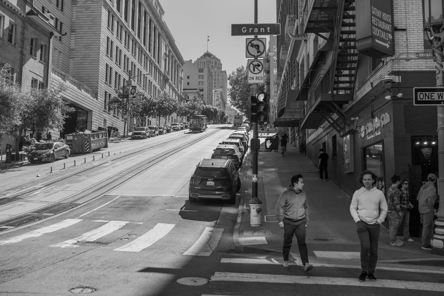 Black and white street scene showing historic architecture along a steep hill in San Francisco with parked cars and pedestrians.